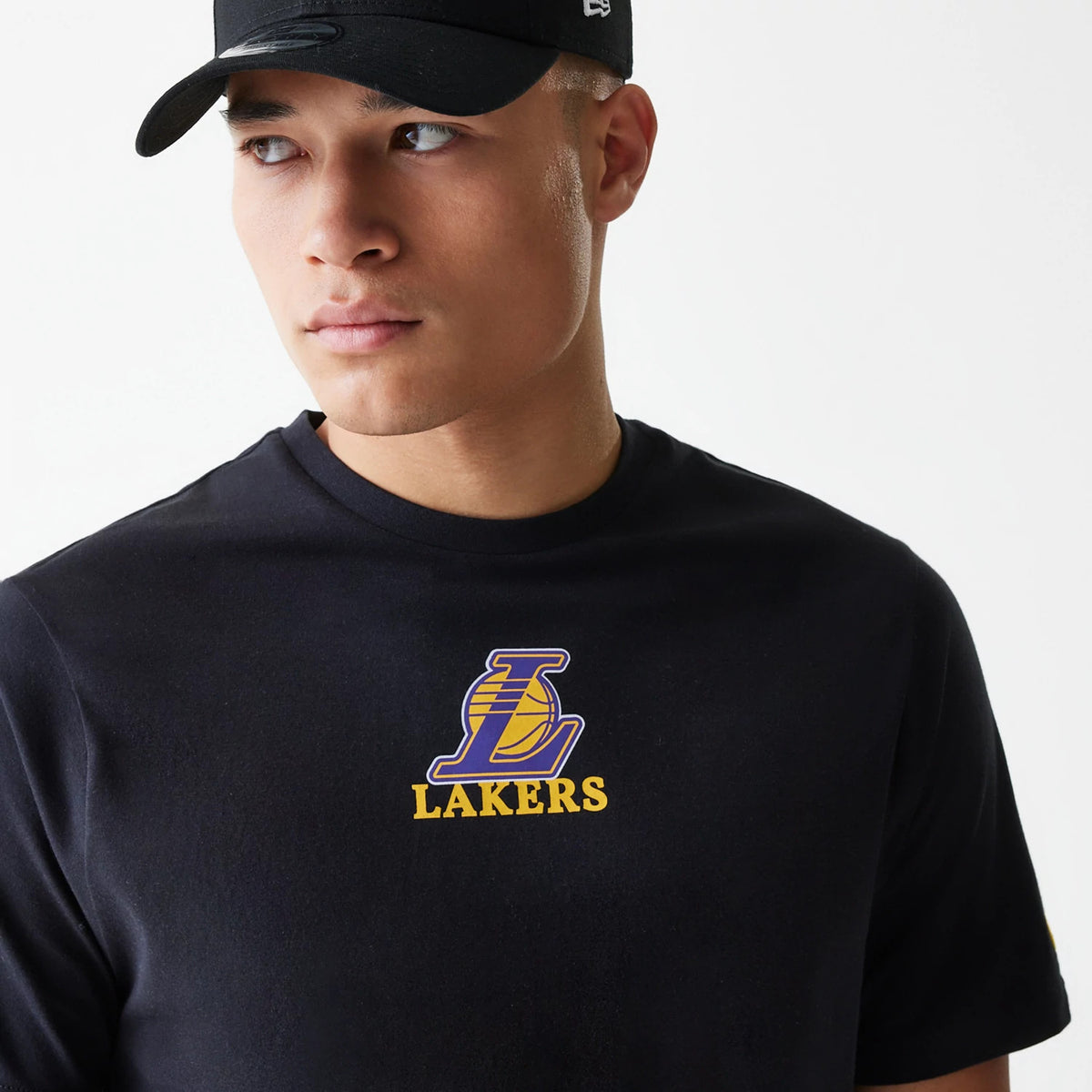 New Era LA Lakers NBA Logo Black T-Shirt, on-model view showing the black T-shirt with a large purple and yellow Lakers logo and debossed lettering on the chest, a black cap with a raised New Era logo worn by the model.