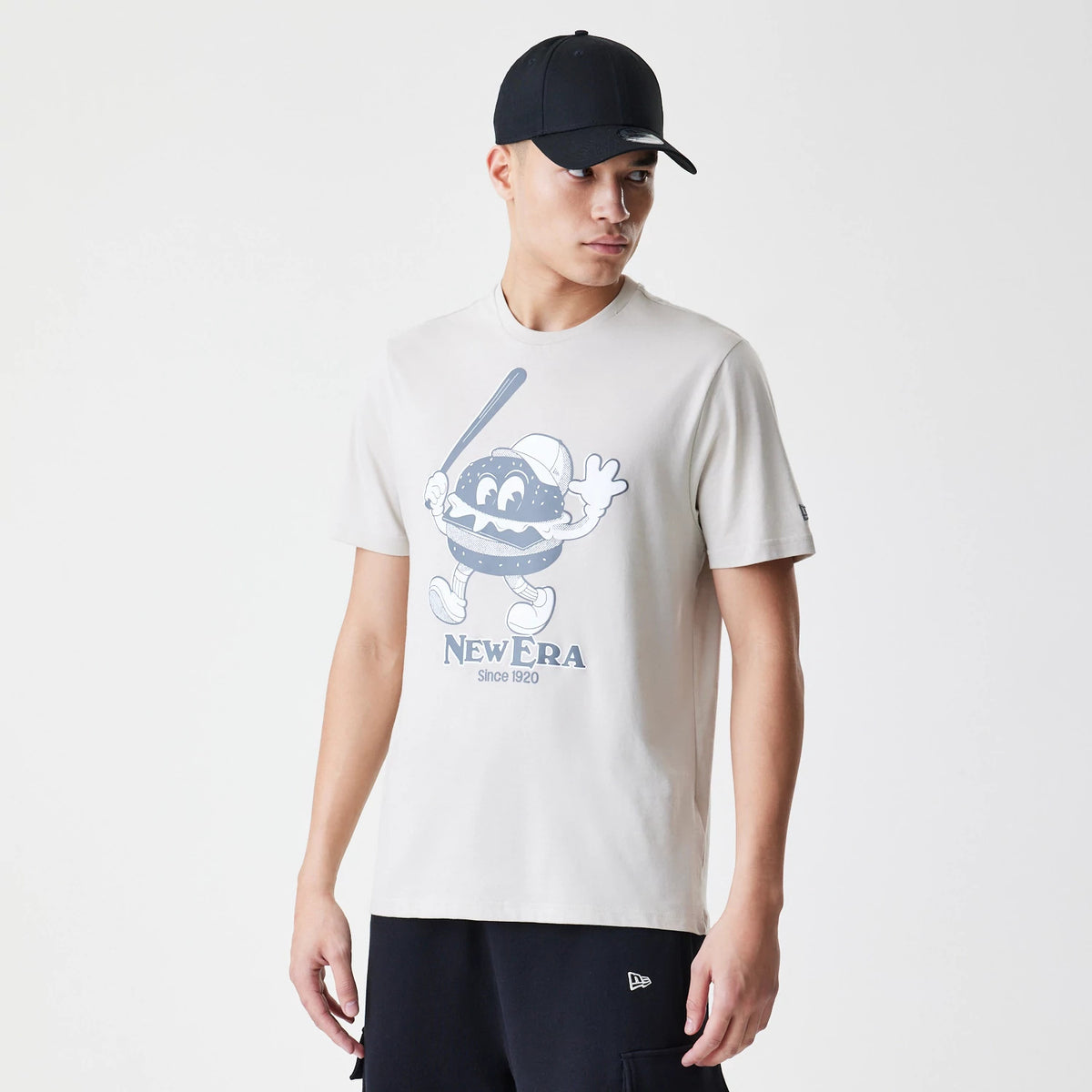 New Era Burger Graphic Cream T-Shirt, on-model view showing the cream shirt with a large cartoon burger graphic on the front, featuring a debossed New Era logo beneath and a man wearing a black cap.