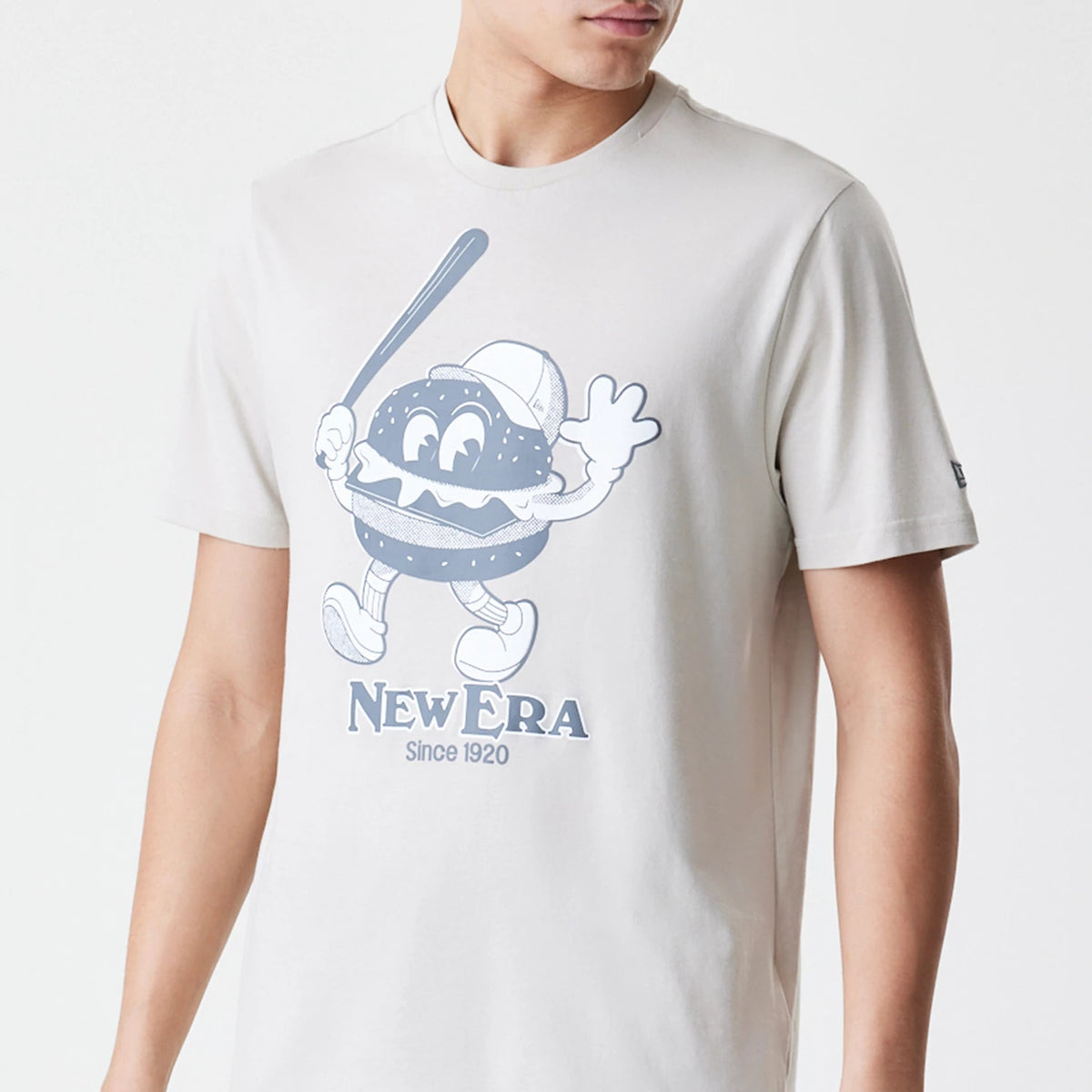 New Era Burger Graphic Cream T-Shirt, front view showing a large playful burger character holding a bat printed in blue on the cream fabric, with 'New Era Since 1920' lettering below and a small debossed New Era logo on the sleeve.