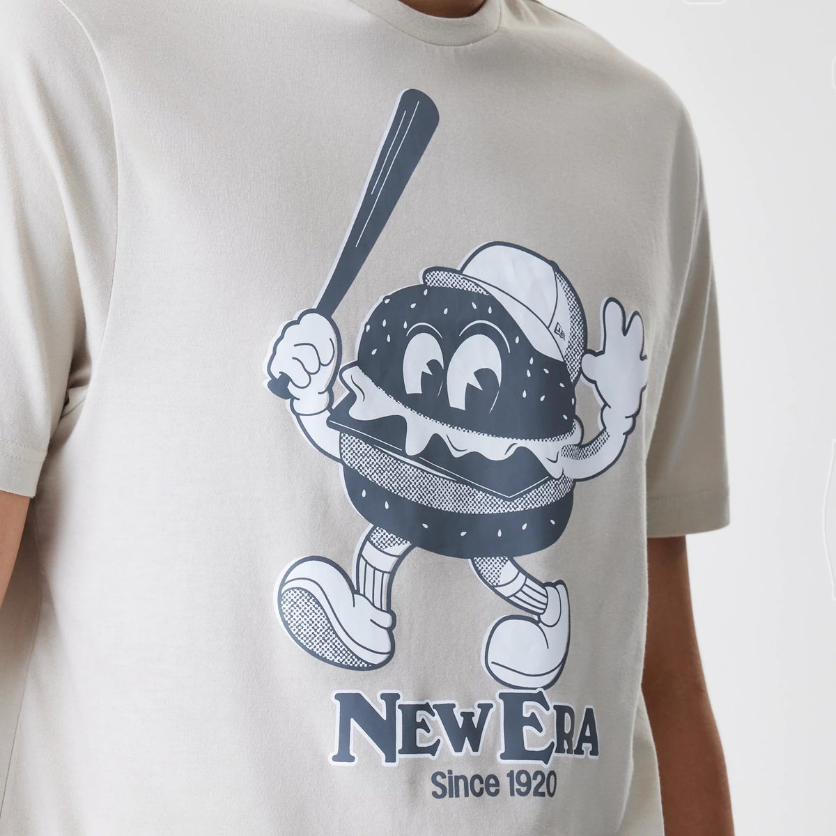 New Era Burger Graphic Cream T-Shirt, front view showing a large printed graphic of a cartoon burger character with eyes, arms, and legs holding a baseball bat, and the New Era logo with Since 1920 text below.