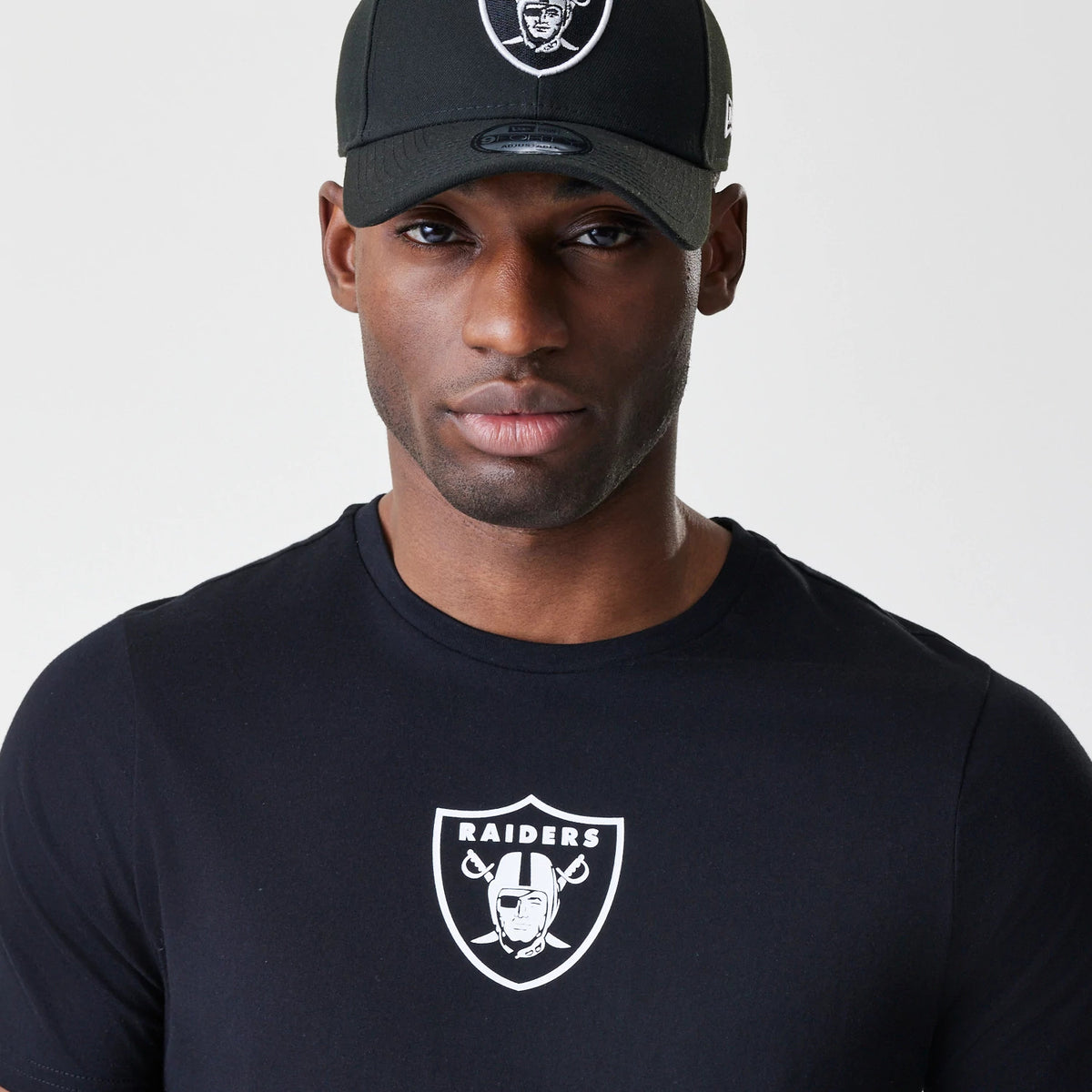 New Era Las Vegas Raiders NFL Logo Black T-Shirt, on-model view showing a large white Raiders logo printed on the chest, worn with a black cap featuring a raised team logo at the front and a small New Era logo on the side.