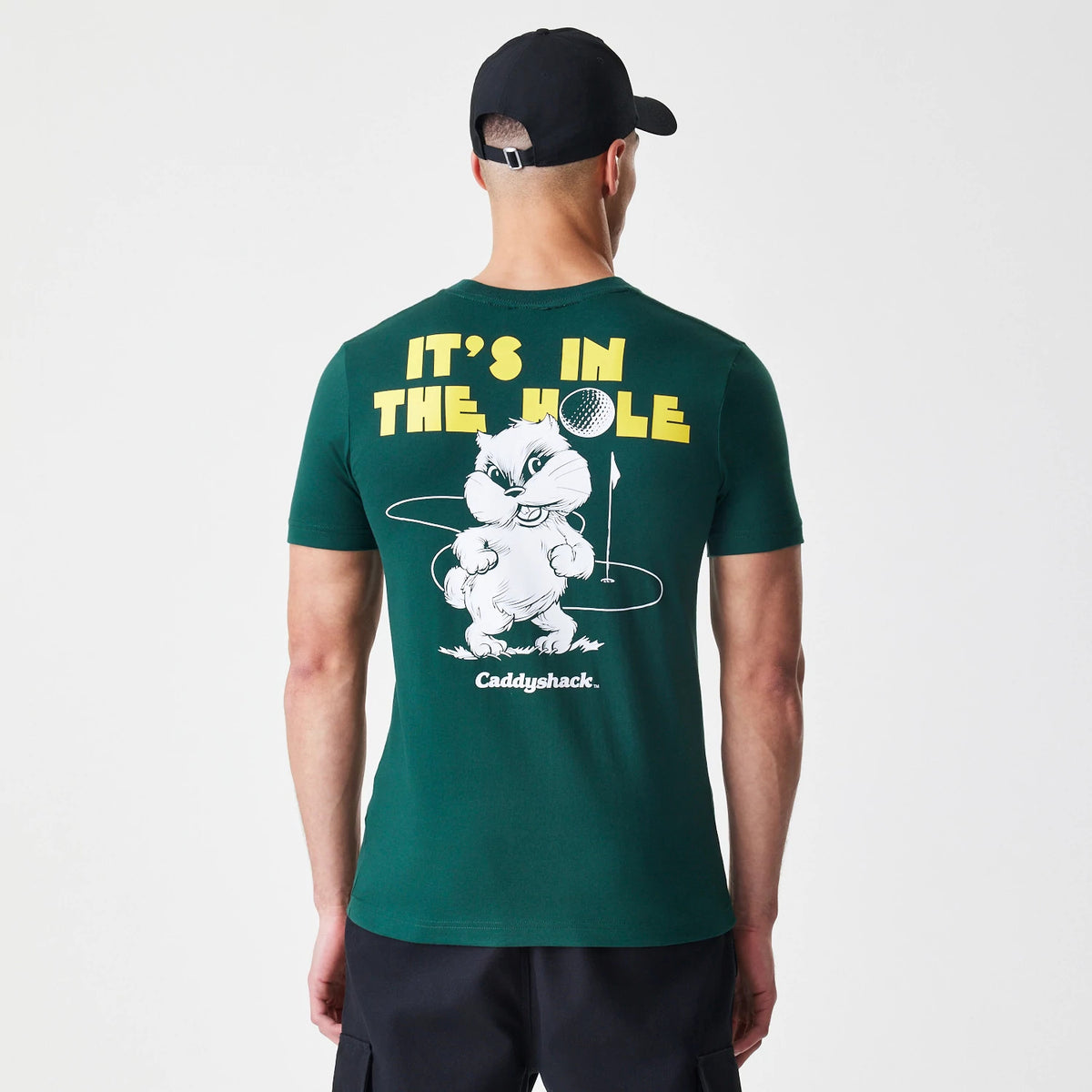 New Era Caddyshack It's In The Hole Dark Green T-Shirt, rear view showing a large white cartoon animal print with a golf ball and yellow text above, worn by a model in black shorts and cap.