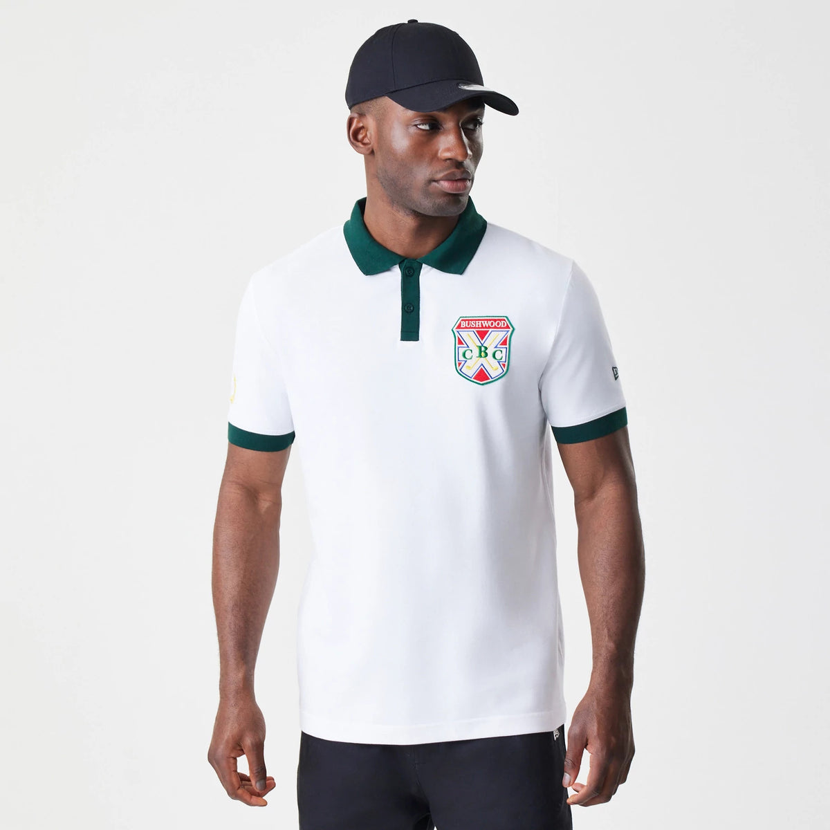 New Era Caddyshack BushWood White Polo T-Shirt, on-model view showing the white polo with a green collar and sleeve trims, a colourful embroidered BushWood CBC crest on the left chest, paired with a black cap on the model's head.
