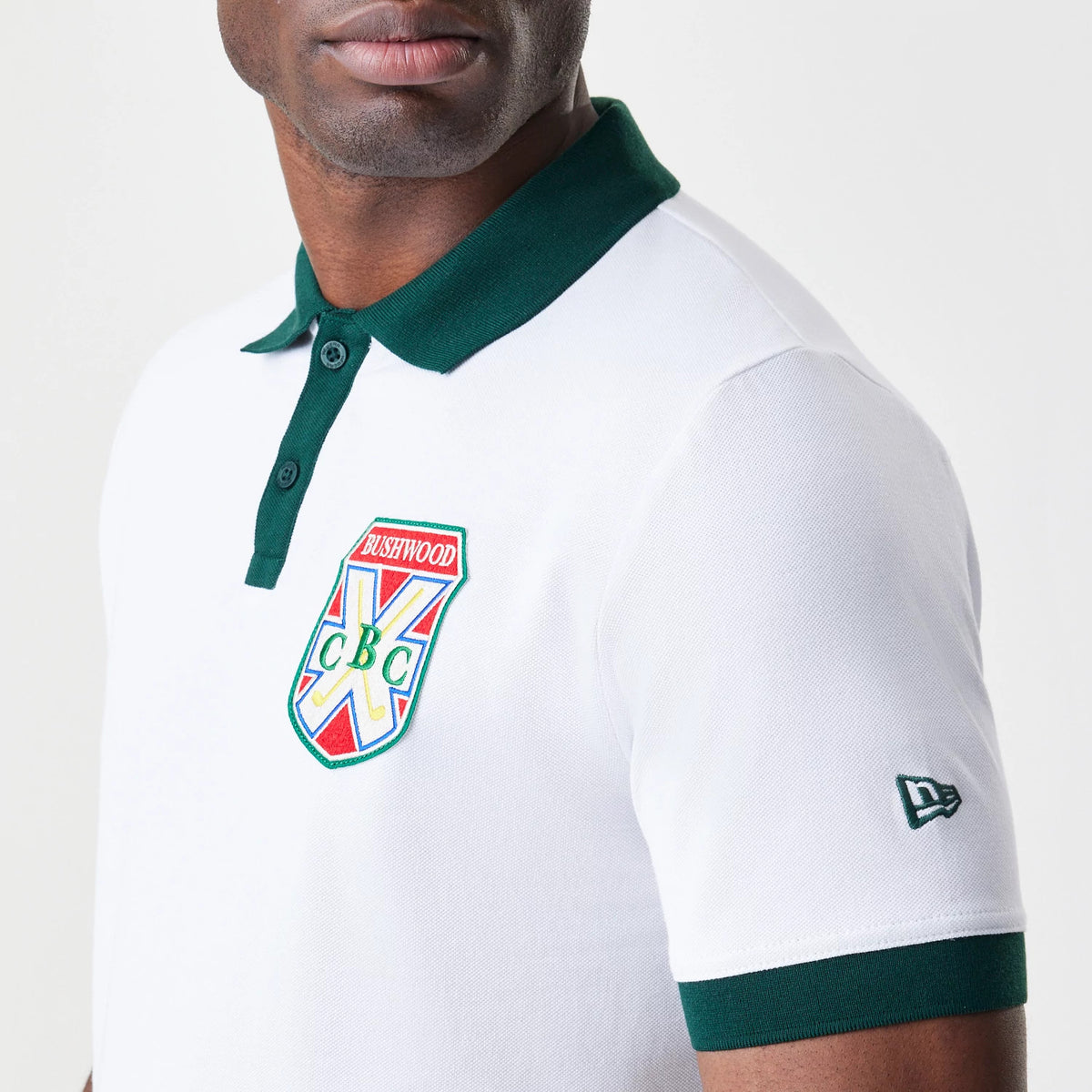 New Era Caddyshack BushWood White Polo T-Shirt, side view showing the white shirt with a green collar and embroidered BushWood crest on the chest, a raised New Era logo on the left sleeve and green edging on the sleeve cuffs.