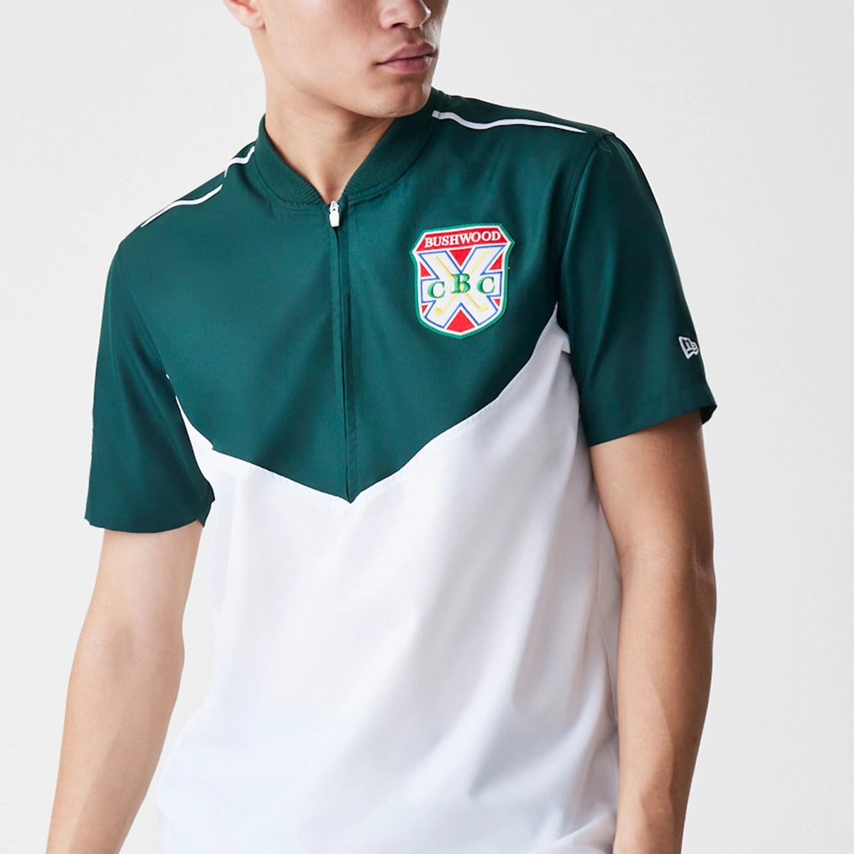New Era Caddyshack BushWood White 1/4 Zip Polo T-Shirt, front view showing a green upper section with a colourful embroidered BushWood CBC logo on the left chest, a quarter zip down the centre, and the New Era logo debossed on the left sleeve.