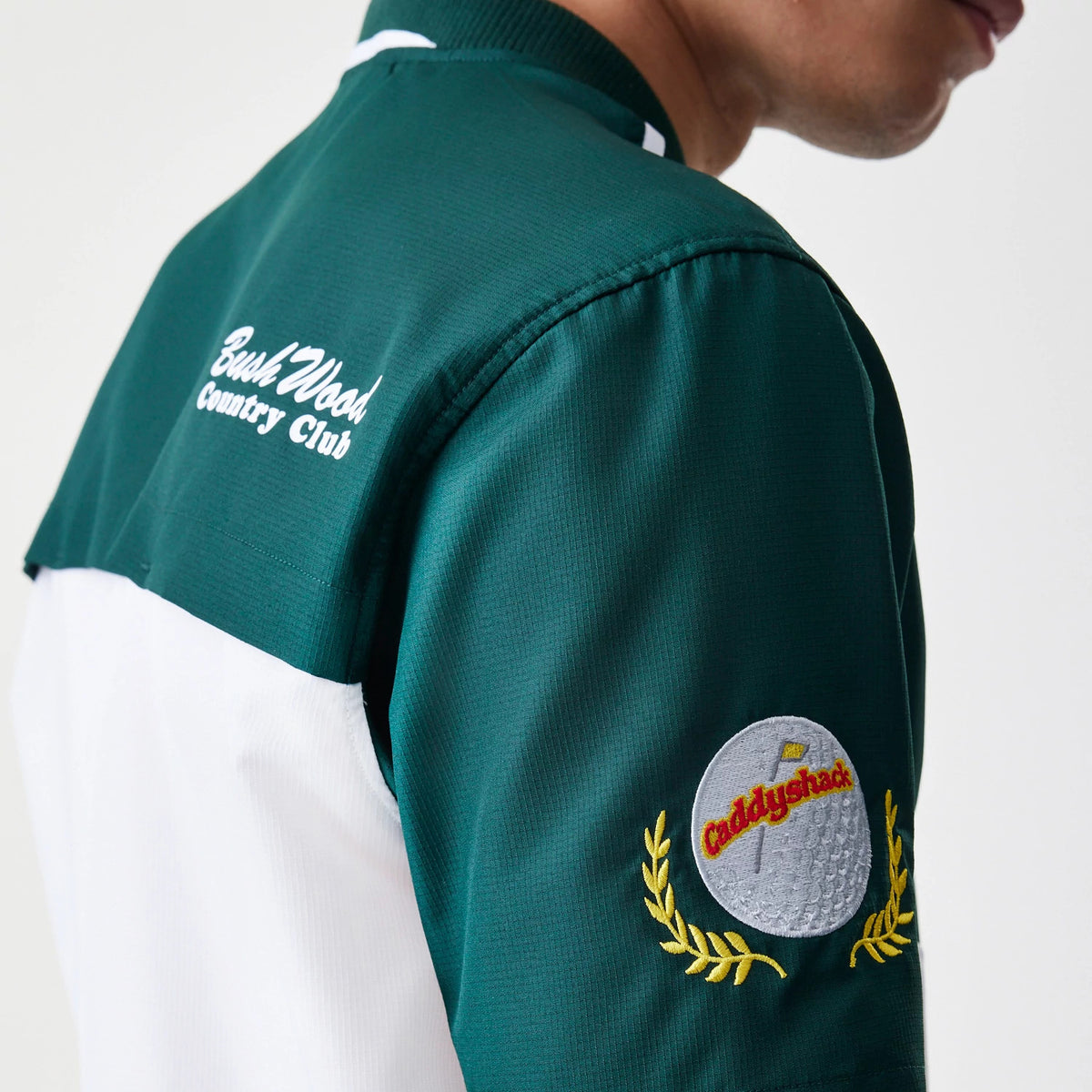 New Era Caddyshack BushWood White 1/4 Zip Polo T-Shirt, three-quarter view showing green sleeves with BushWood Country Club in white cursive on the back and an embroidered Caddyshack logo with a golf motif and yellow laurels on the upper arm.