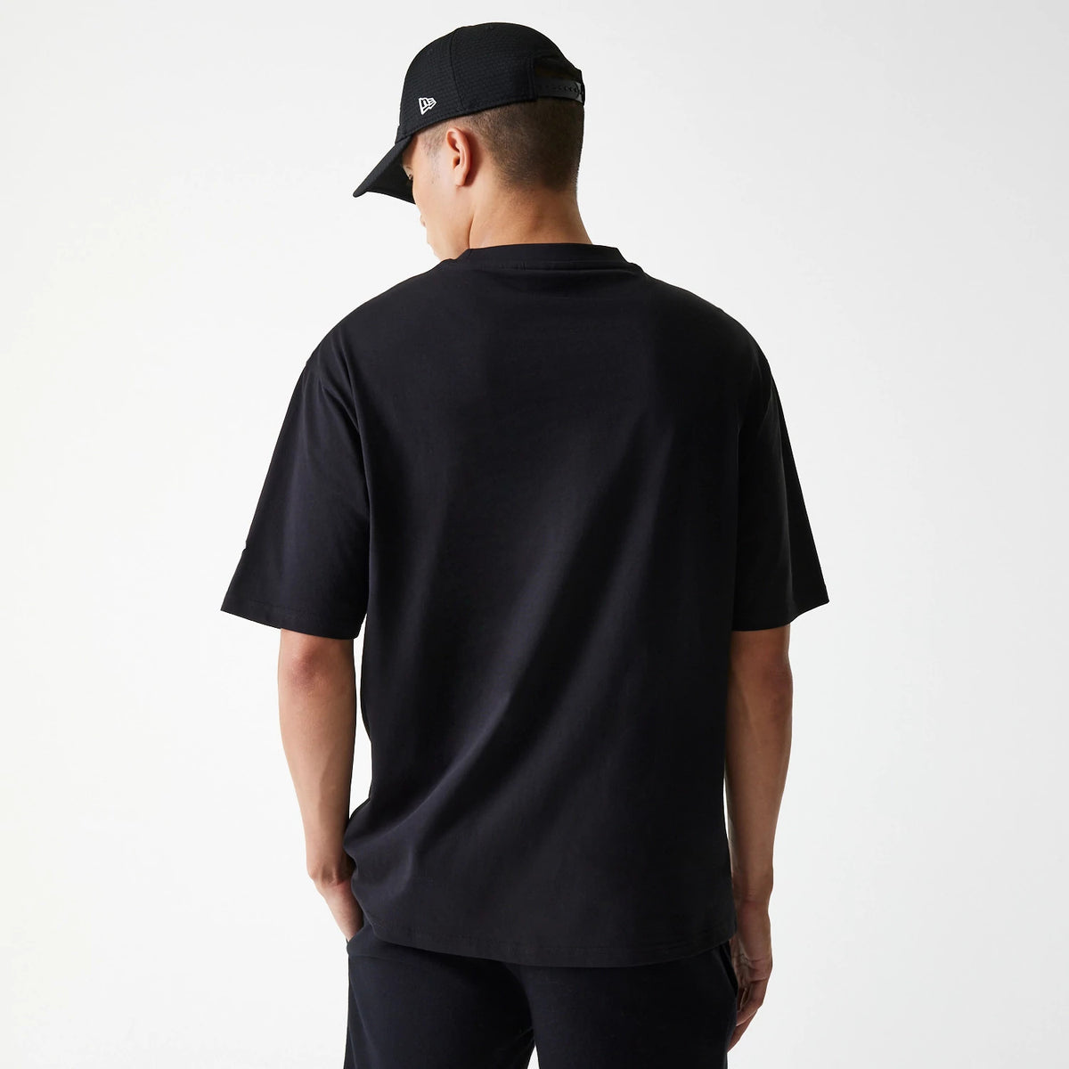 New Era New York Yankees MLB League Essential Black Oversized T-Shirt, back view showing the plain black fabric with a loose fit, short sleeves, worn by a person also wearing a black cap displaying a raised New Era logo on the side.