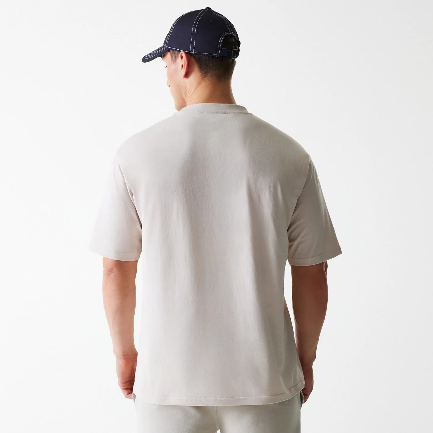 New Era New York Yankees MLB League Essential Cream Oversized T-Shirt, rear view showing the plain cream fabric with short sleeves and a relaxed fit, worn by a model with a navy cap featuring white stitching and an adjustable back strap.