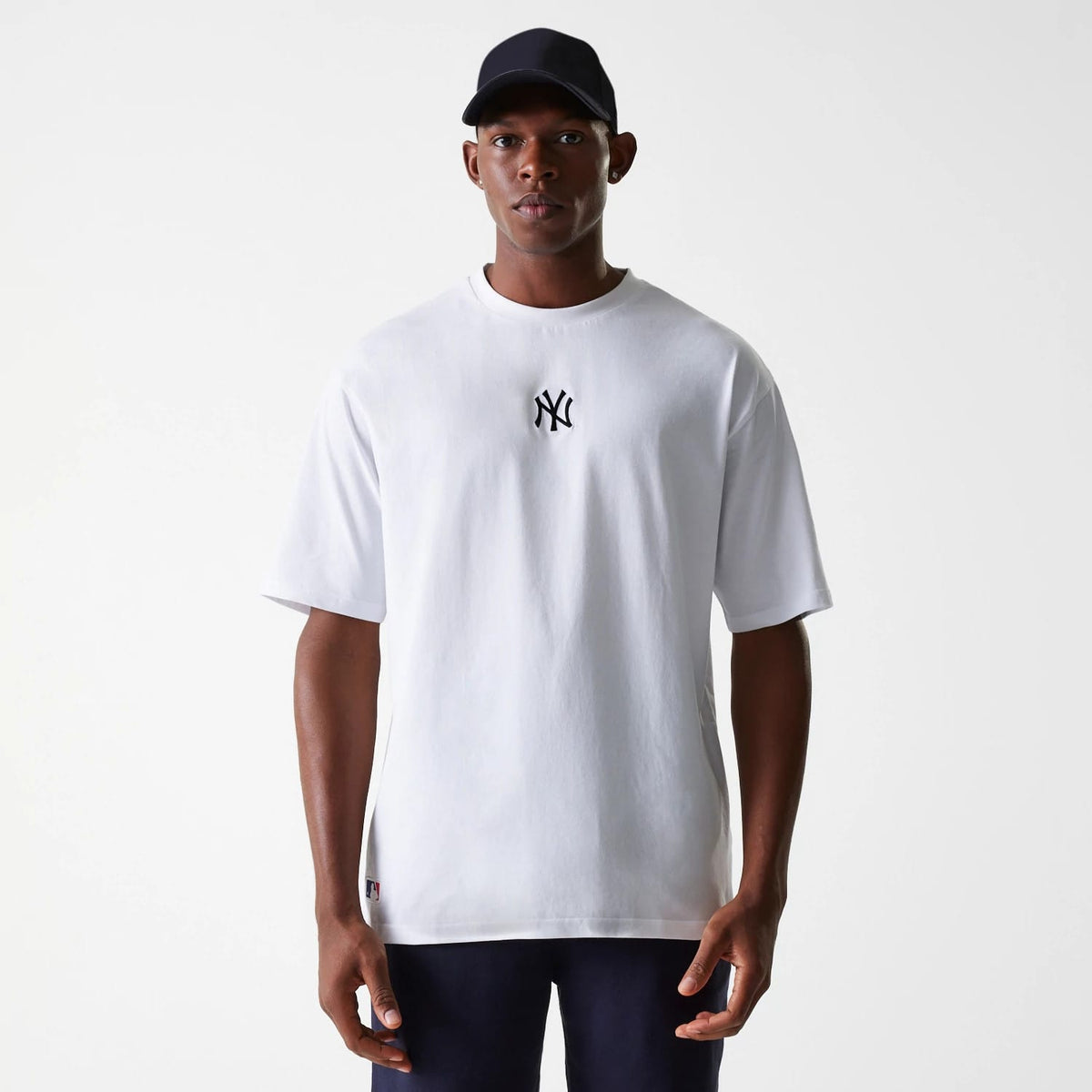 New Era New York Yankees MLB League Essential White Oversized T-Shirt, front view showing a black embroidered New York Yankees logo centred on the chest and a small label sewn at the hem, worn by a model in a dark cap.