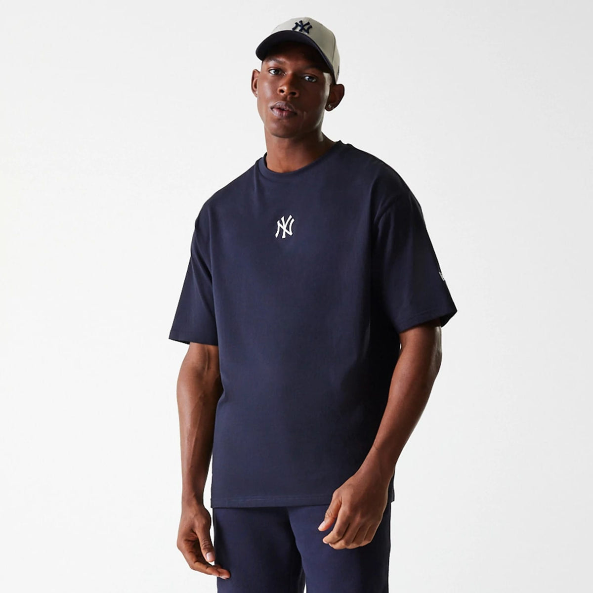 New Era New York Yankees MLB League Essential Navy Oversized T-Shirt, on-model view showing a navy oversized fit with a white embroidered Yankees logo on the chest, worn with navy trousers and a beige cap with a navy Yankees logo.