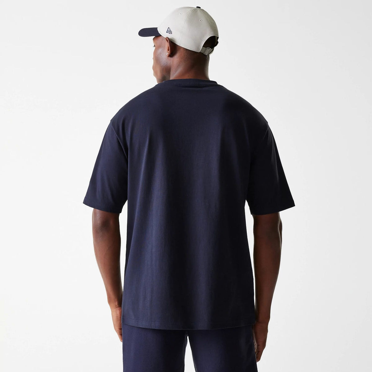 New Era New York Yankees MLB League Essential Navy Oversized T-Shirt, rear view showing the plain navy back of the oversized T-shirt worn by a model, paired with a light-coloured cap having the New Era debossed logo on the side.