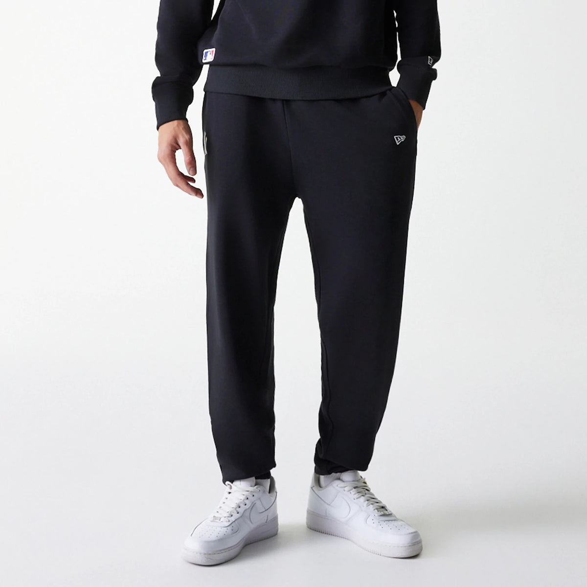 New Era New York Yankees MLB League Essential Midi French Terry Black Joggers, front view on a model with white trainers, showing the New Era flag logo embroidered on the left thigh and an adjustable waistband with side pockets.