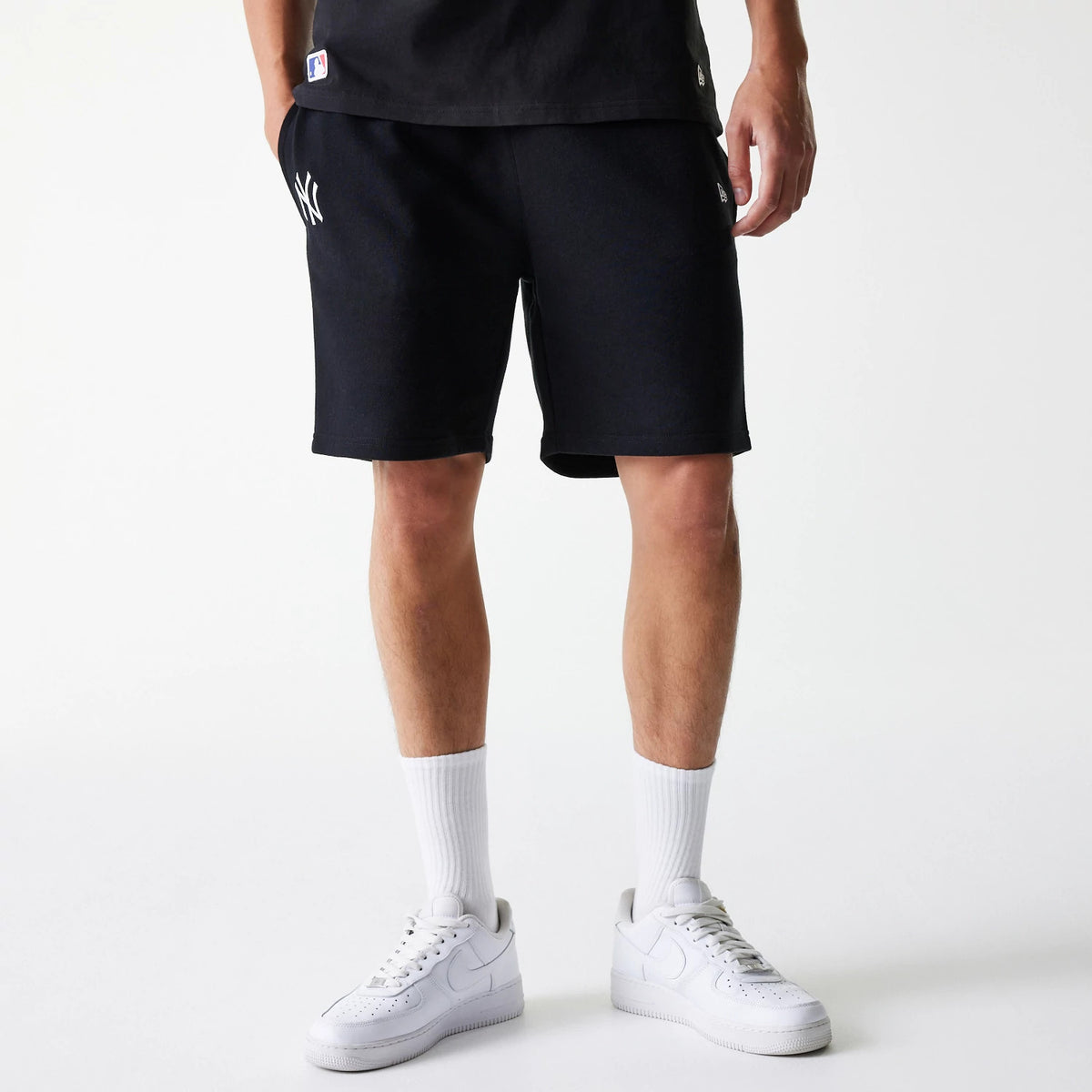 New Era New York Yankees MLB League Essential French Terry Black Shorts, on-model view showing the raised white New York Yankees logo on the left thigh, above knee length cut, paired with white socks and trainers.
