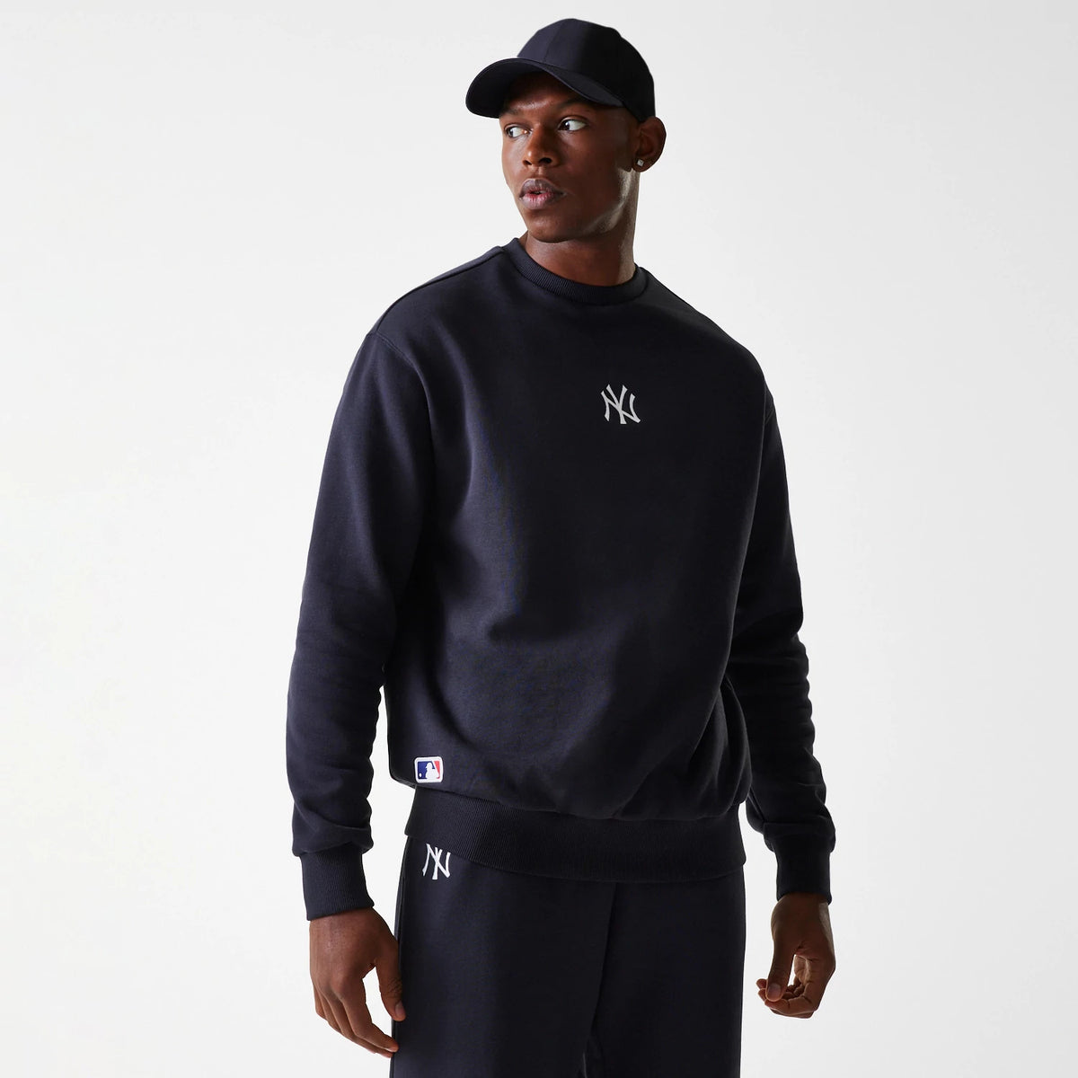 New Era New York Yankees MLB League Essential French Terry Navy Crew Neck Sweater, on a model wearing matching navy sweatpants and cap, featuring a white NY logo on the chest and an MLB Batterman patch near the waistband.