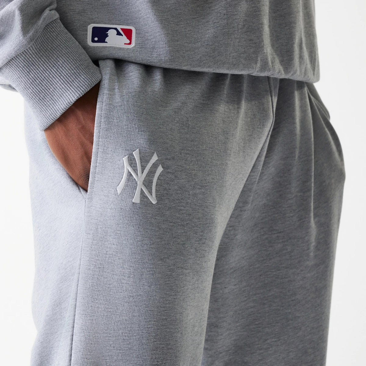 New Era New York Yankees MLB League Essential Midi French Terry Grey Joggers, side view showing the white embroidered NY logo on the left thigh and a red, white, and blue MLB logo on the upper right pocket area.