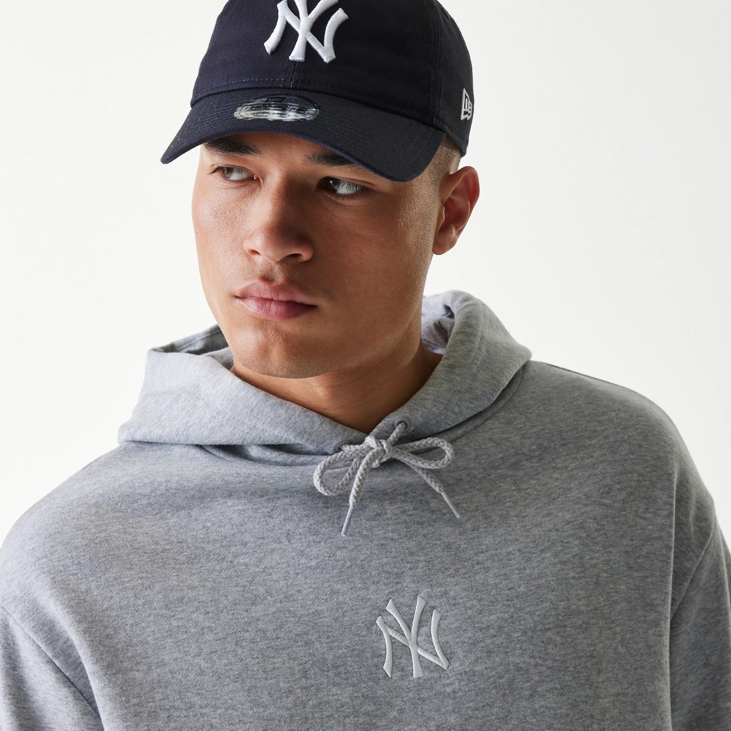 New Era New York Yankees MLB League Essential Grey Oversized Pullover Hoodie, on-model view showing a light grey hoodie with a raised New York Yankees logo on the chest, model wearing a navy cap with a white raised logo.