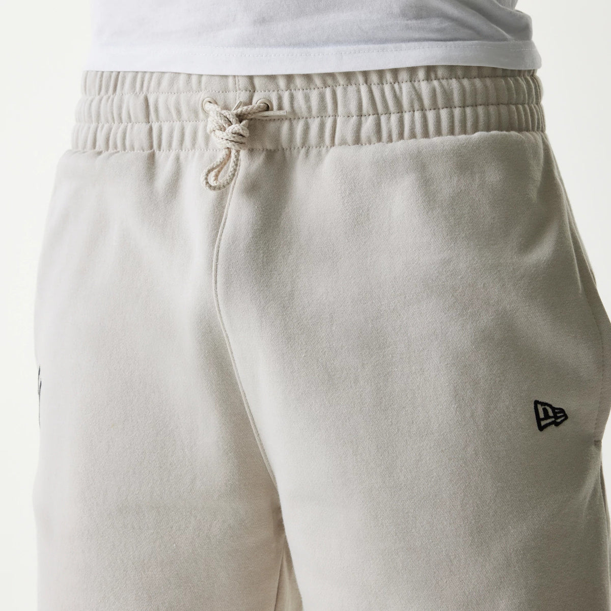 New Era New York Yankees MLB League Essential Midi Cream Shorts, front view showing the cream fabric with an elasticated waistband and drawstring, a raised black New Era logo on the left thigh, and stitching details along the seams.