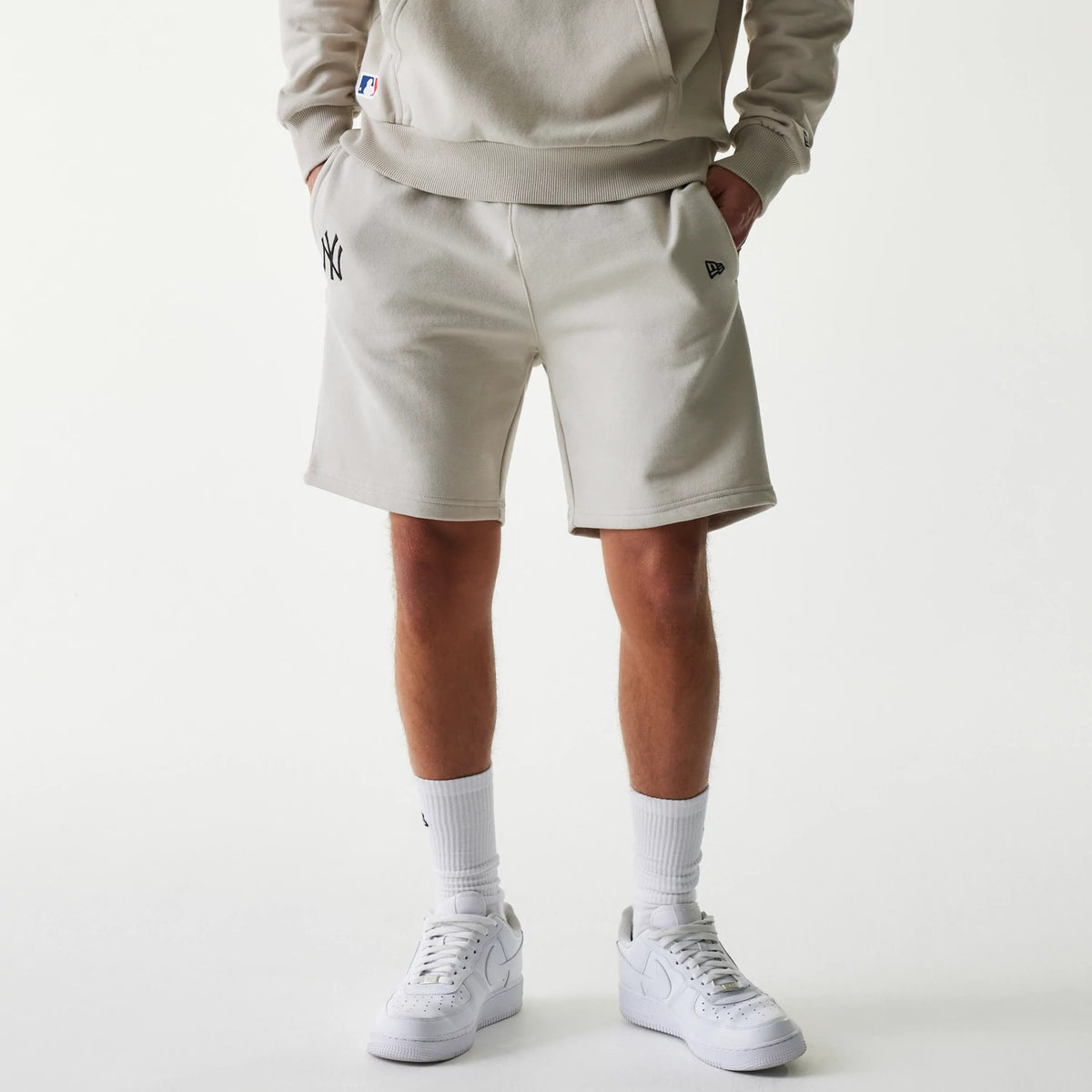 New Era New York Yankees MLB League Essential Midi Cream Shorts, on-model view showing cream shorts with an embroidered New York Yankees logo on the left thigh and a debossed New Era logo on the right thigh, worn with white trainers and socks.