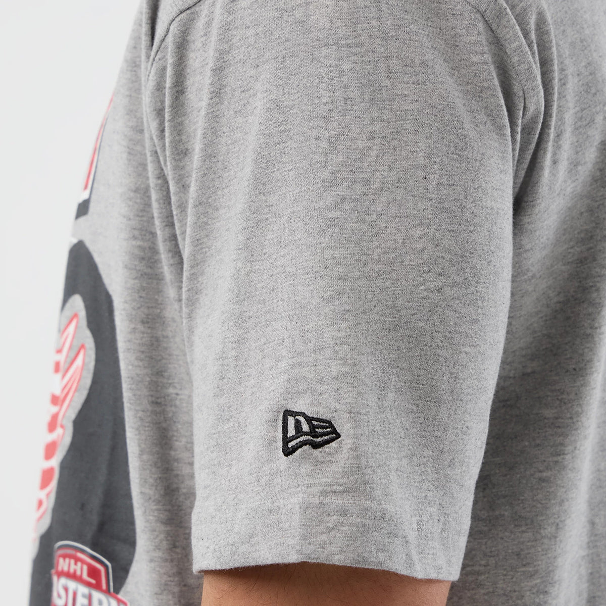 New Era Detroit Red Wings NHL Sport Classic Grey T-Shirt, side view showing the short sleeve with an embroidered New Era logo in black near the hem and part of the graphic design on the torso in black, red, and white.