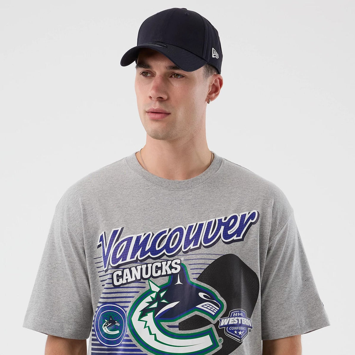 New Era Vancouver Canucks NHL Sport Classic Grey T-Shirt, on-model view showing a large team logo and name printed across the chest in blue and green, paired with a navy cap featuring a raised New Era logo on the side.