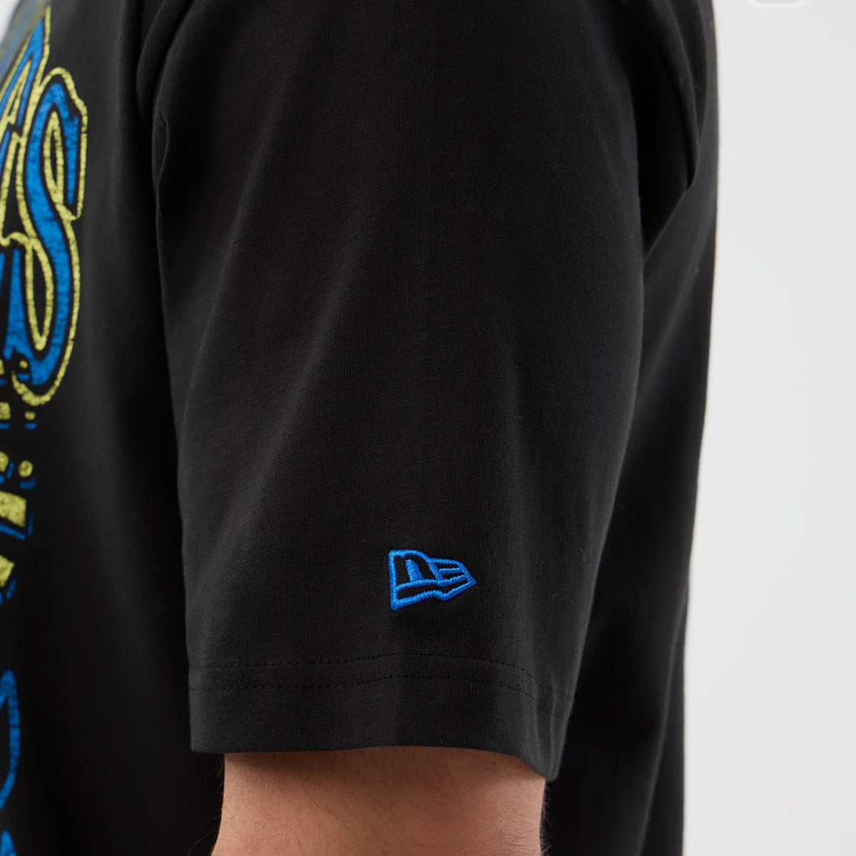 New Era LA Dodgers MLB Player Life Graphic Black T-Shirt, side view showing the short sleeve with a raised blue New Era logo embroidered near the cuff and part of a large blue and yellow graphic design extending from the sleeve.