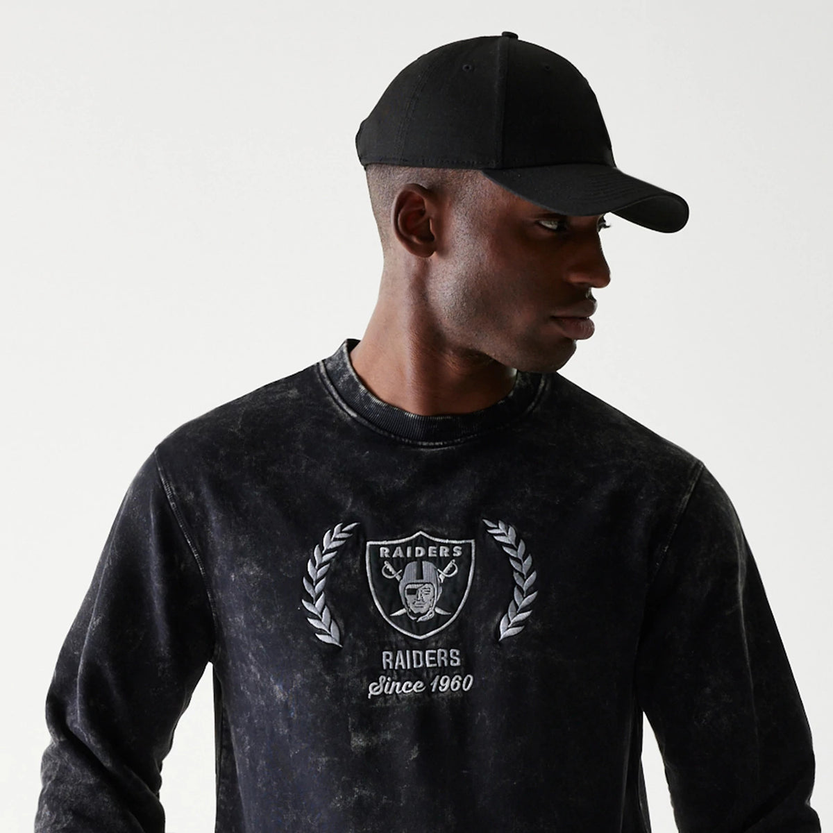 New Era Las Vegas Raiders NFL Throwback Black Crew Neck Sweater, on-model showing the embroidered Raiders shield logo and laurel wreath on the chest, paired with a plain black cap, styled with a distressed wash effect.