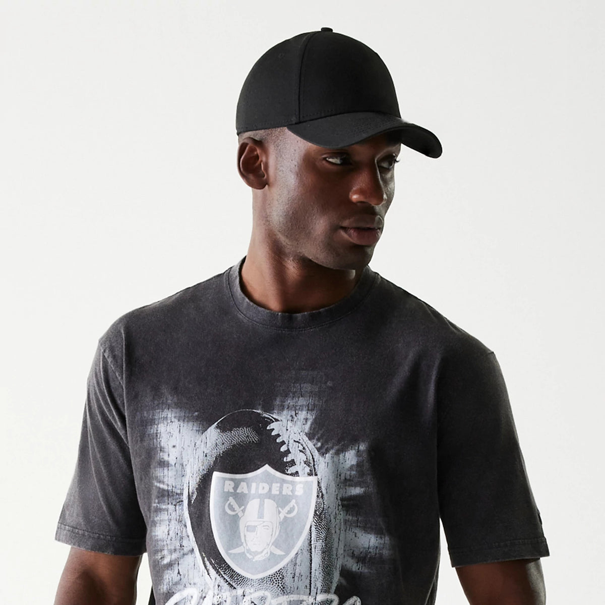 New Era Las Vegas Raiders NFL Sport Night Black T-Shirt, on-model view showing the black and grey T-shirt with a large faded football and Raiders logo on the chest, worn with a plain black cap against a light background.