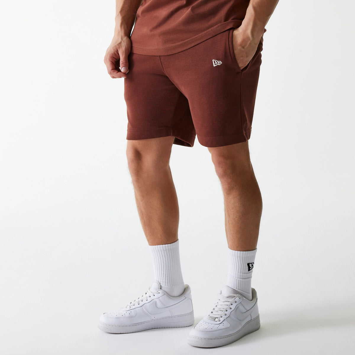 New Era New York Yankees MLB Midi League Essential Brown Shorts, on-model view showing knee-length brown shorts with a small raised New Era logo on the left thigh, hands in side pockets and paired with white socks and trainers.