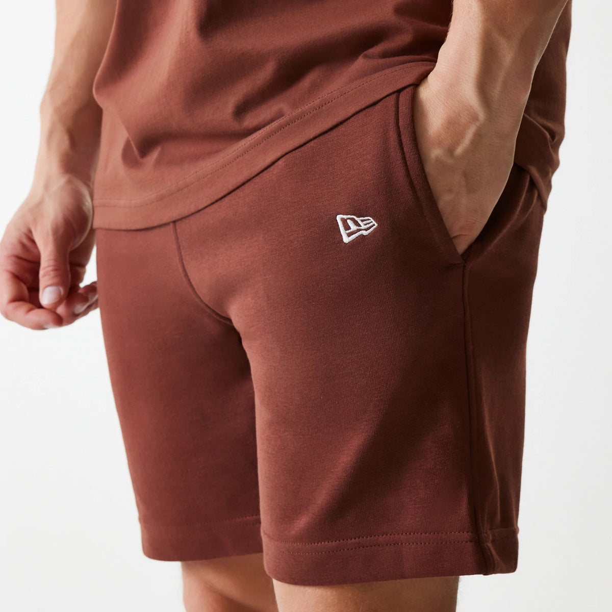 New Era New York Yankees MLB Midi League Essential Brown Shorts, on-model view showing the rich brown fabric with a white embroidered New Era logo on the left thigh, the hand in the pocket and the casual fit around the leg.