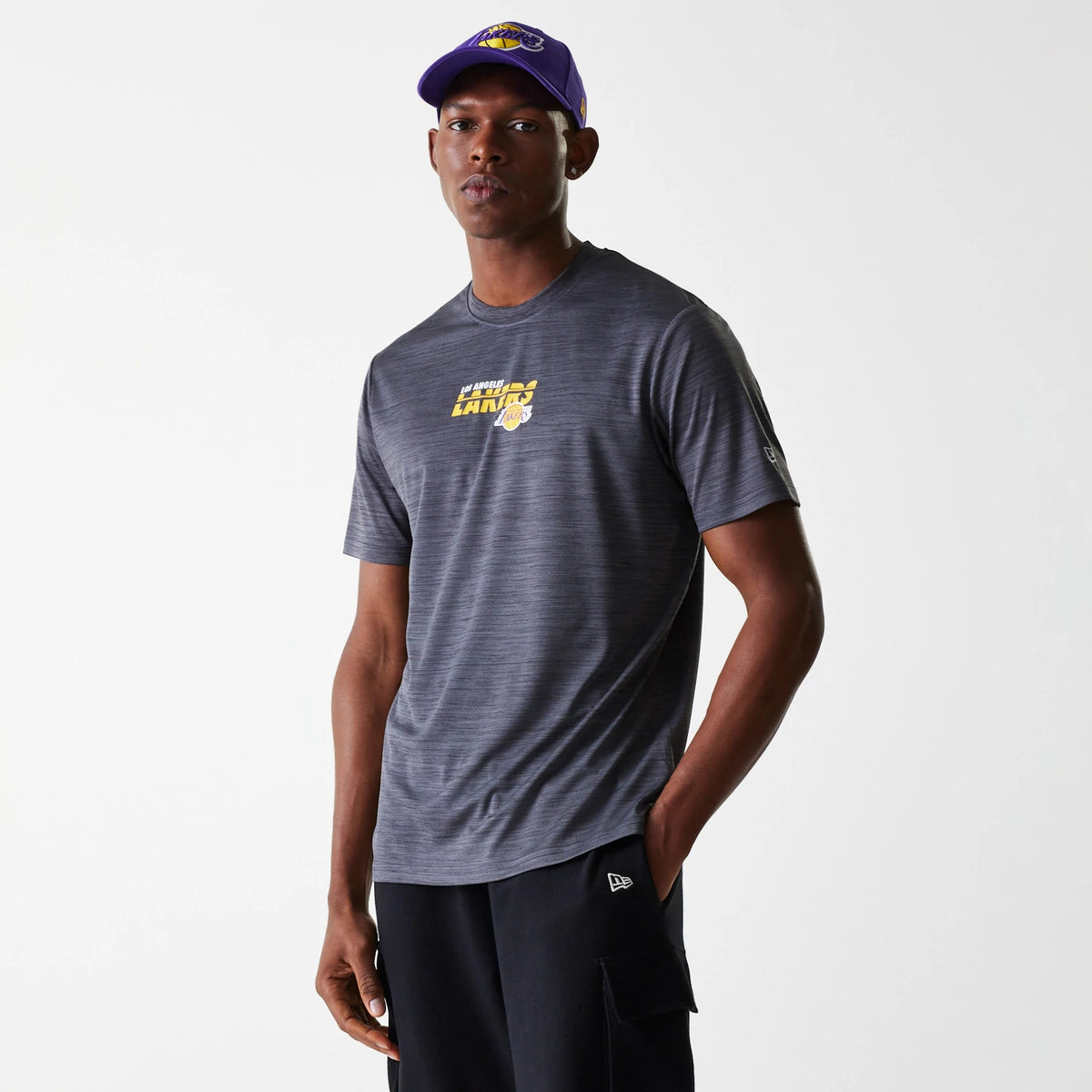 New Era LA Lakers NBA Essentials Black T-Shirt, on-model view showing the grey short-sleeve design with a raised yellow LA Lakers logo centred on the chest, a New Era logo on the left sleeve and a purple cap on the model's head.