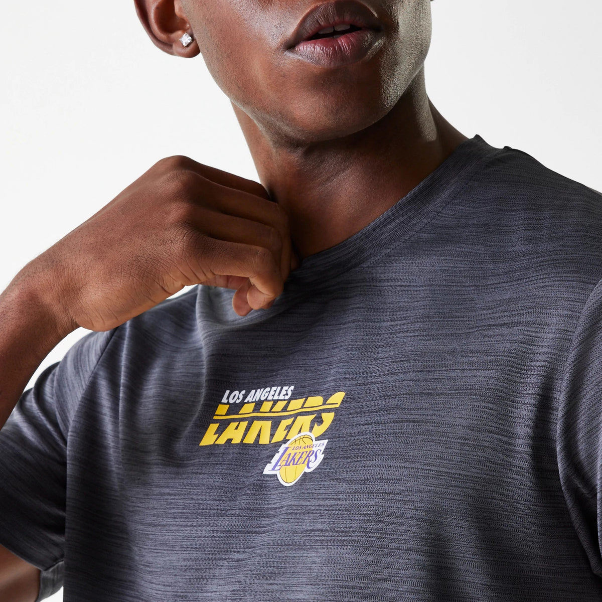 New Era LA Lakers NBA Essentials Black T-Shirt, on-model view showing the black t-shirt with a Los Angeles Lakers logo printed in yellow and white across the chest, worn by a person holding the collar.