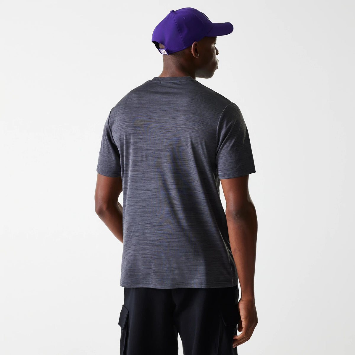 New Era LA Lakers NBA Essentials Black T-Shirt, rear view showing the plain black fabric with short sleeves, worn by a model in black trousers, a raised white NBA logo on a purple cap matching the outfit.