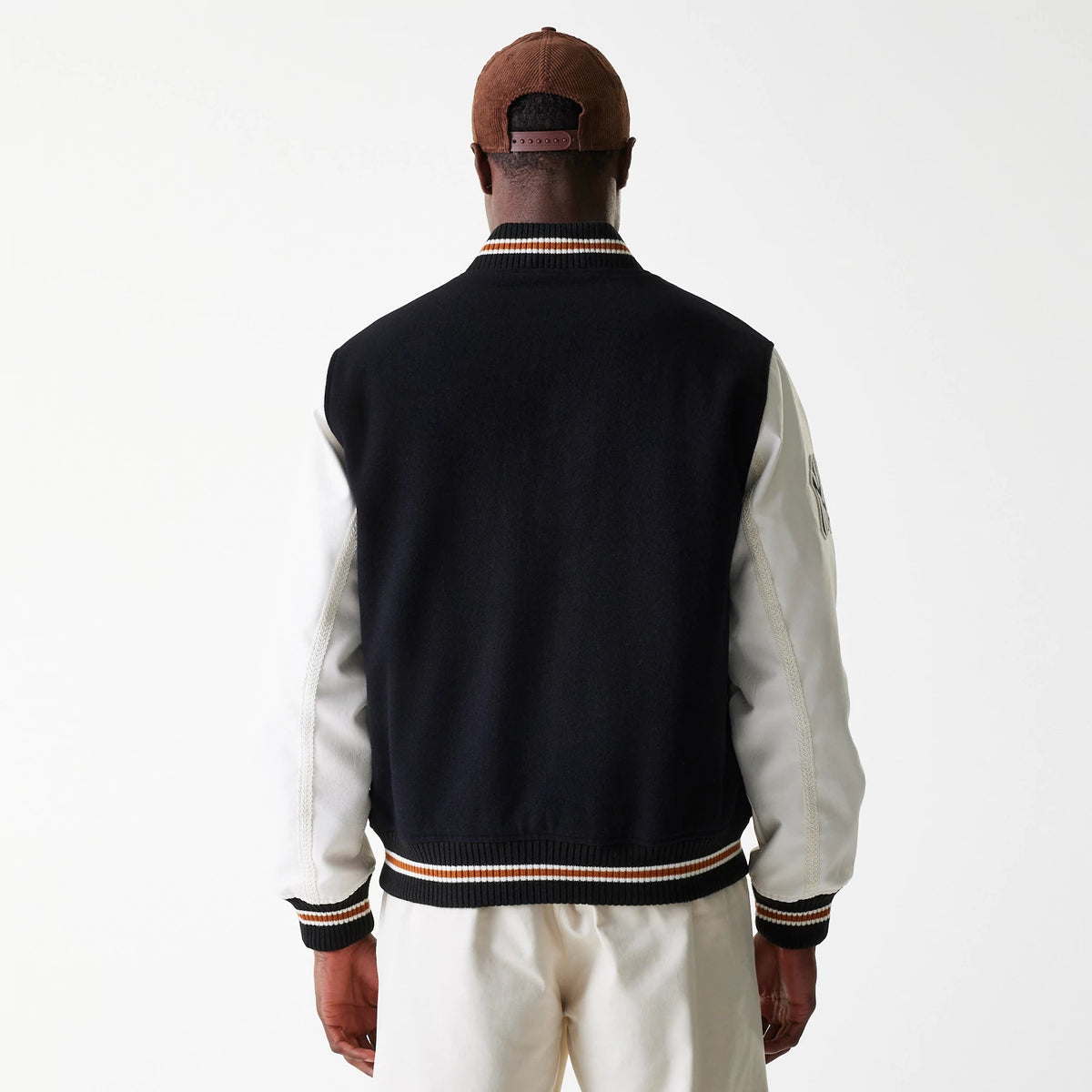 New Era New York Yankees MLB Wordmark Black Varsity Jacket, rear view showing the black back and white sleeves with striped ribbed hem and cuffs, worn by a person in a brown cap facing away.
