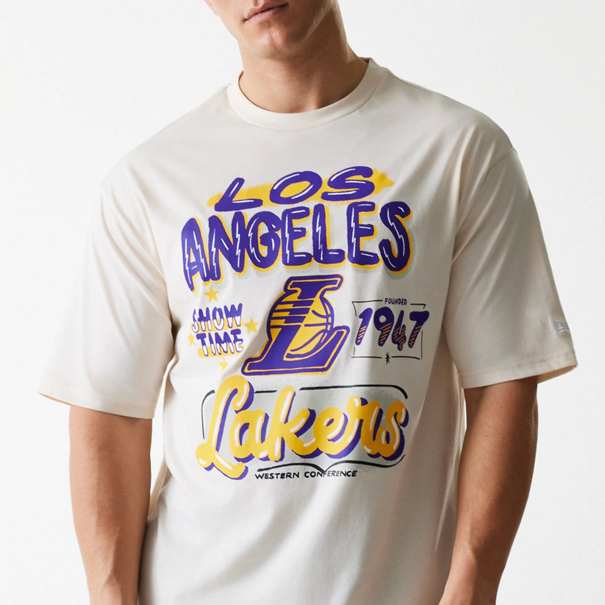 New Era LA Lakers NBA Graphic Light Beige Oversized T-Shirt, front view showing the bold purple and yellow Los Angeles Lakers graphic centred on the chest, with a series of text and logo elements in an artistic style.
