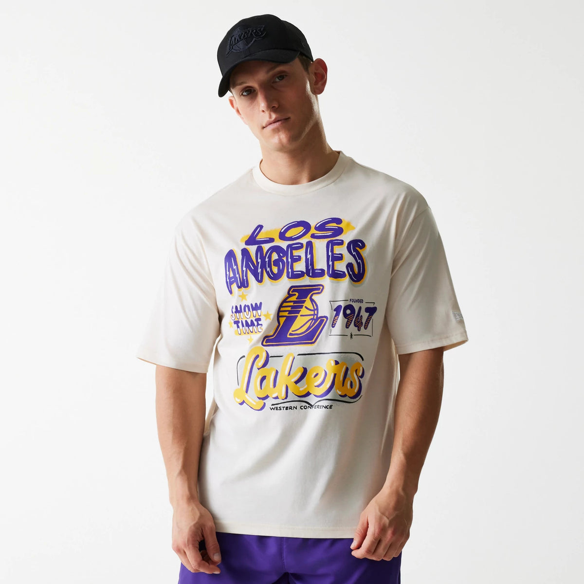 New Era LA Lakers NBA Graphic Light Beige Oversized T-Shirt, on-model view showing the large purple and gold graphic text across the front with Lakers prominently displayed, paired with a black cap featuring a raised Lakers logo.