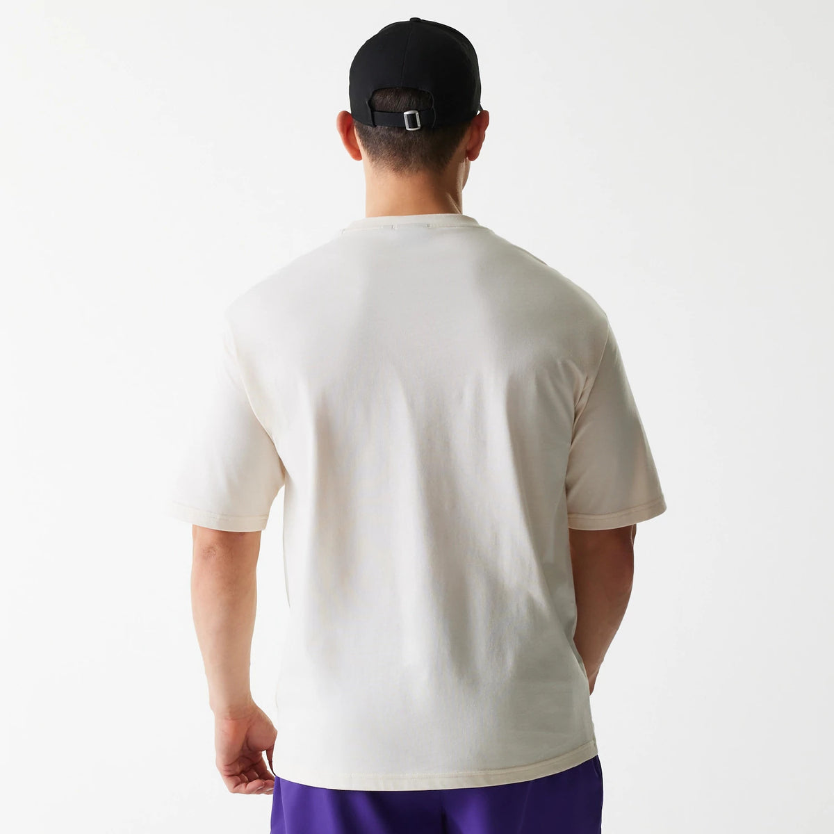 New Era LA Lakers NBA Graphic Light Beige Oversized T-Shirt, rear view showing the plain light beige back with a loose fit and short sleeves, worn by a person with a black cap and purple shorts.