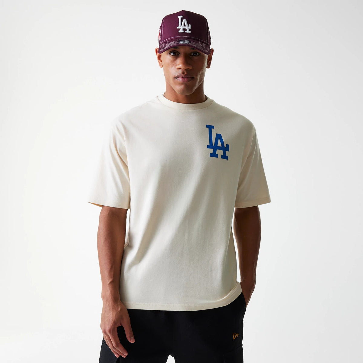New Era LA Dodgers MLB Baseball Graphic Light Beige Oversized T-Shirt, front view featuring a blue LA logo on the left chest, shown on-model with a burgundy cap displaying a raised white LA logo on the crown.