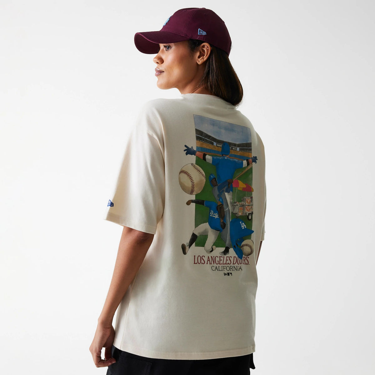 New Era LA Dodgers MLB Baseball Graphic Light Beige Oversized T-Shirt, on-model view showing the back graphic of a baseball scene in vivid colours, with a small New Era logo embroidered on the sleeve and a purple cap worn by the model.