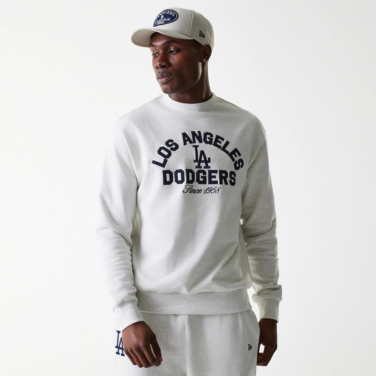 New Era LA Dodgers MLB Heritage Pastel Grey Oversized Crew Neck Sweater, on a model styled with matching sweatpants, featuring navy Los Angeles Dodgers lettering across the chest and a small New Era flag logo on the left leg.