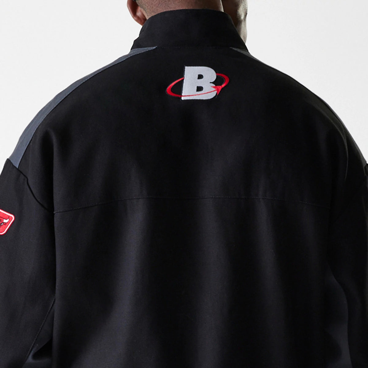 New Era Chicago Bulls NBA Black Motorsports Jacket, rear view showing a raised B logo with a red swoosh centred on the upper back, grey panels on the shoulders, and a small red Chicago Bulls emblem on the left sleeve.