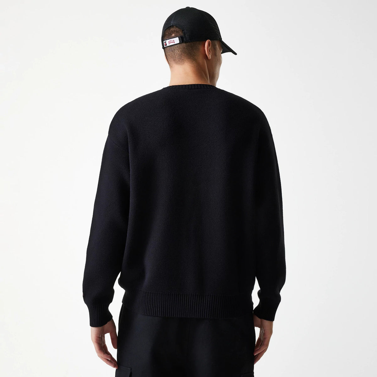 New Era Chicago Bulls NBA Knit Black Oversized Knit Sweater, rear view on a model with a black cap, showing the loose fit of the sweater and ribbed hem and cuffs, highlighting casual style with Chicago Bulls branding on the cap.