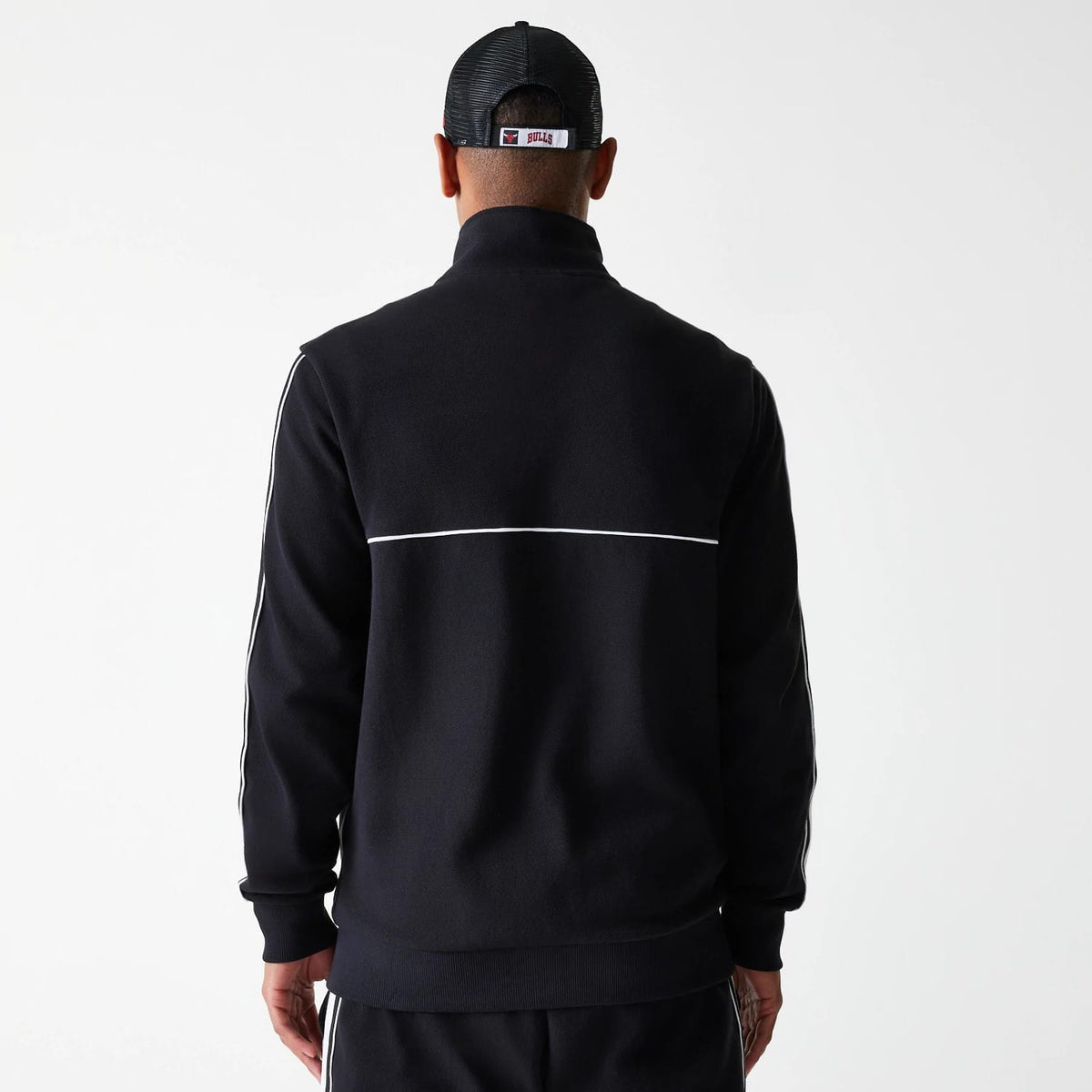 New Era Chicago Bulls NBA Outline Black Track Jacket, back view showing the black fabric with a single white stripe across the upper back and two white stripes along each sleeve, worn by a model wearing a black cap.