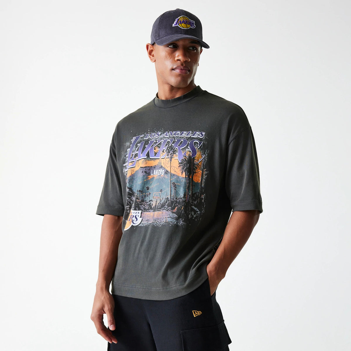 New Era LA Lakers NBA Washed Graphic Grey T-Shirt, on-model view showing the large Lakers graphic on the front, worn by a person in black shorts with a large pocket featuring a raised New Era logo and a grey cap with a Lakers logo.