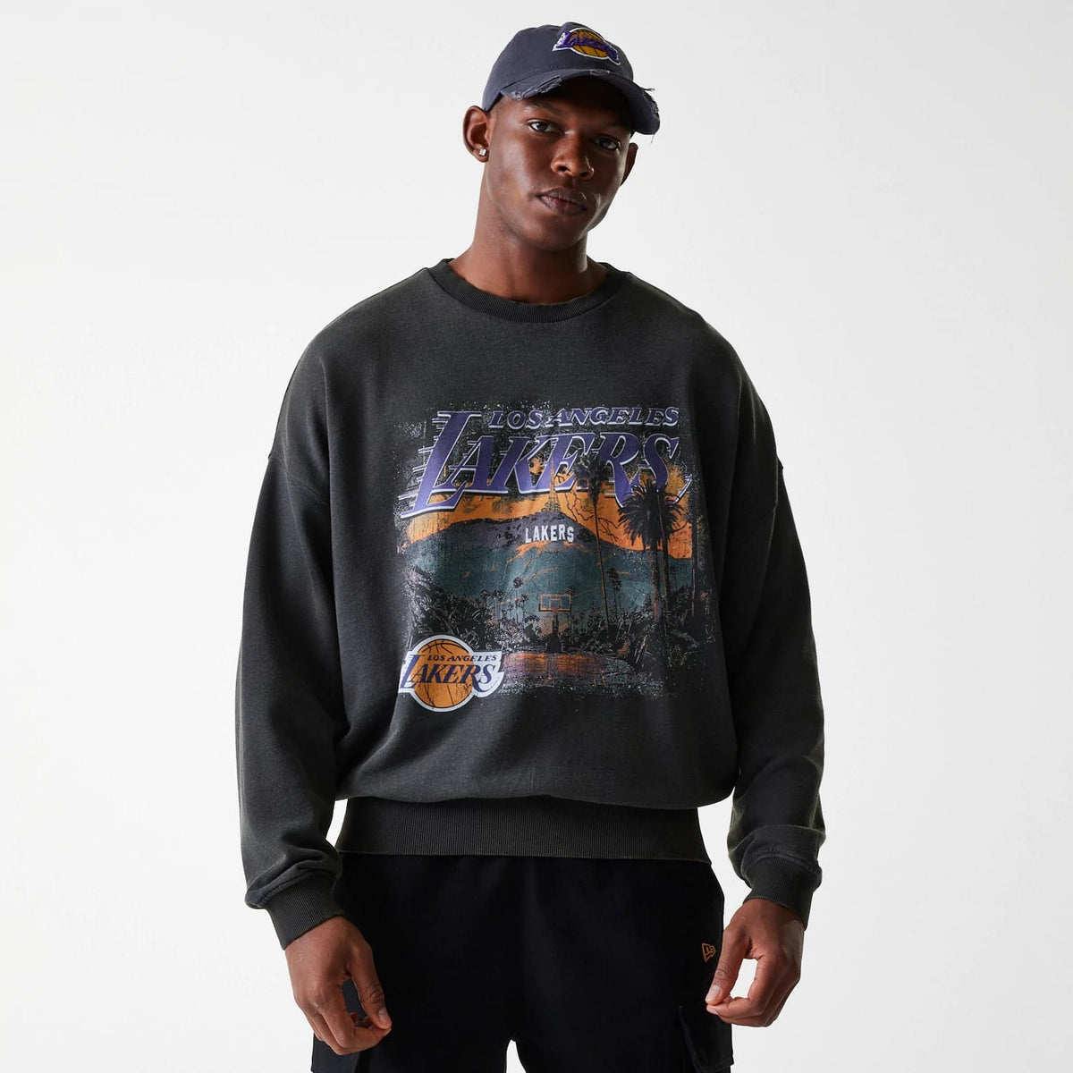 New Era LA Lakers NBA Washed Graphic Grey Oversized Crew Neck Sweater, on-model view with a basketball scene and Lakers logo on the front, contrasted against black trousers, the model wearing a matching cap with Lakers logo.