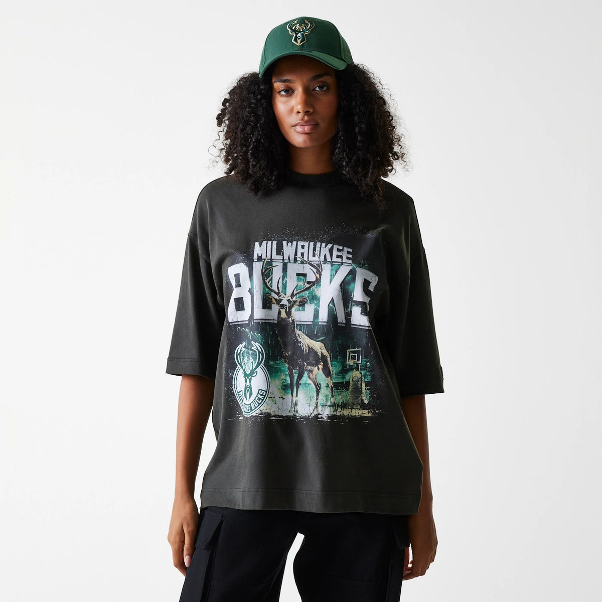 New Era Milwaukee Bucks NBA Washed Graphic Grey T-Shirt, front view showing a large graphic print of the Bucks' logo and a deer in the centre on a washed grey fabric, worn by a model paired with a green cap featuring a raised team logo.