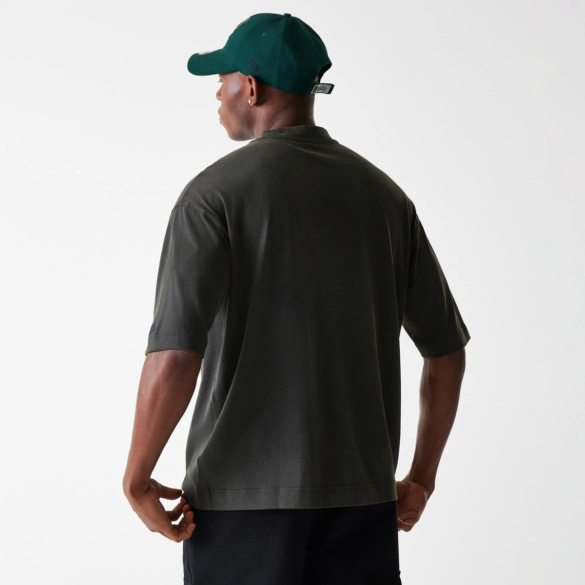 New Era Milwaukee Bucks NBA Washed Graphic Grey T-Shirt, on-model view showing the back of the grey T-shirt with short sleeves, a green cap on the model's head featuring an embossed logo on the side.