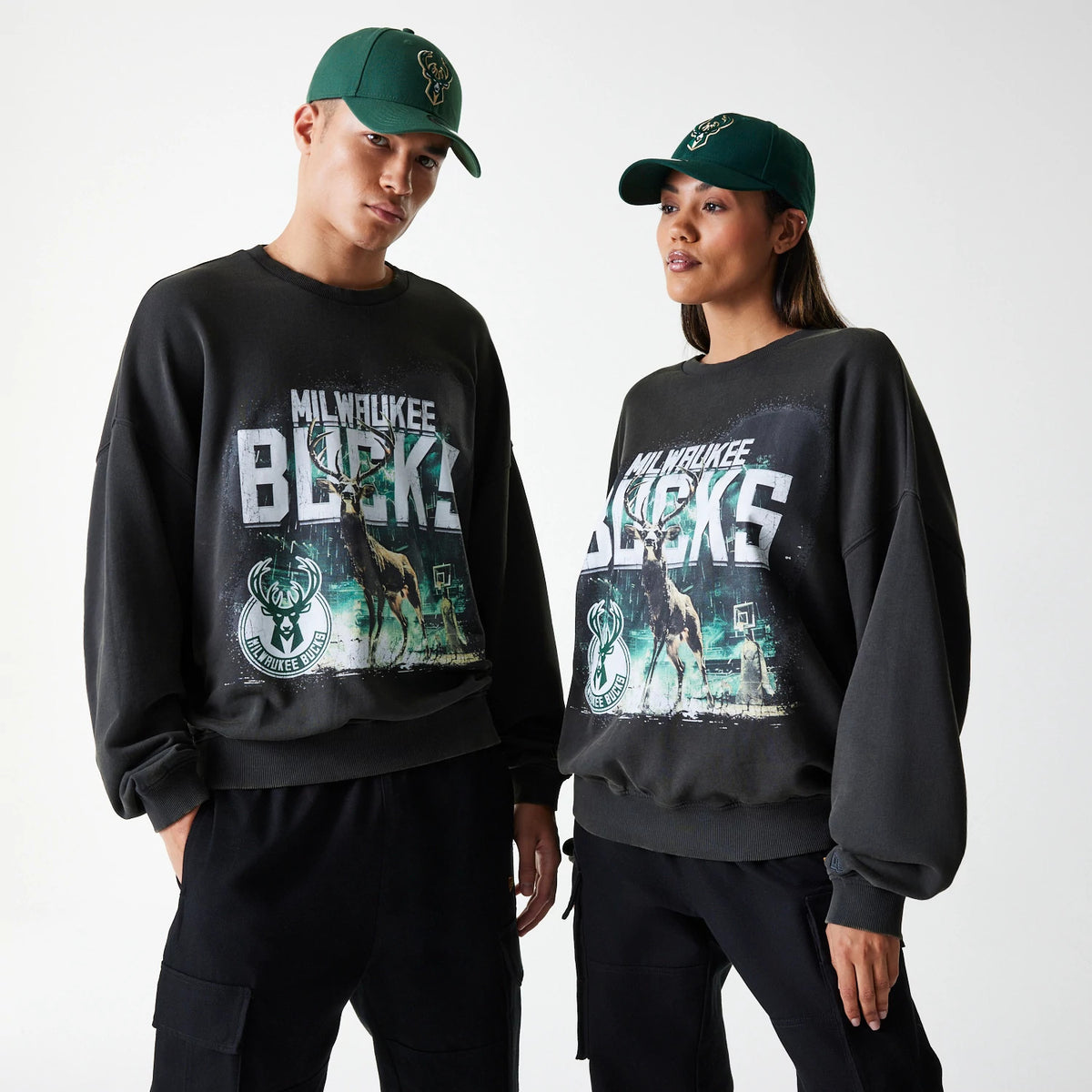 New Era Milwaukee Bucks NBA Washed Graphic Grey Oversized Crew Neck Sweater, front view on a male and female model wearing green Bucks caps, showcasing the large distressed Bucks lettering and mascot graphics on the chest.