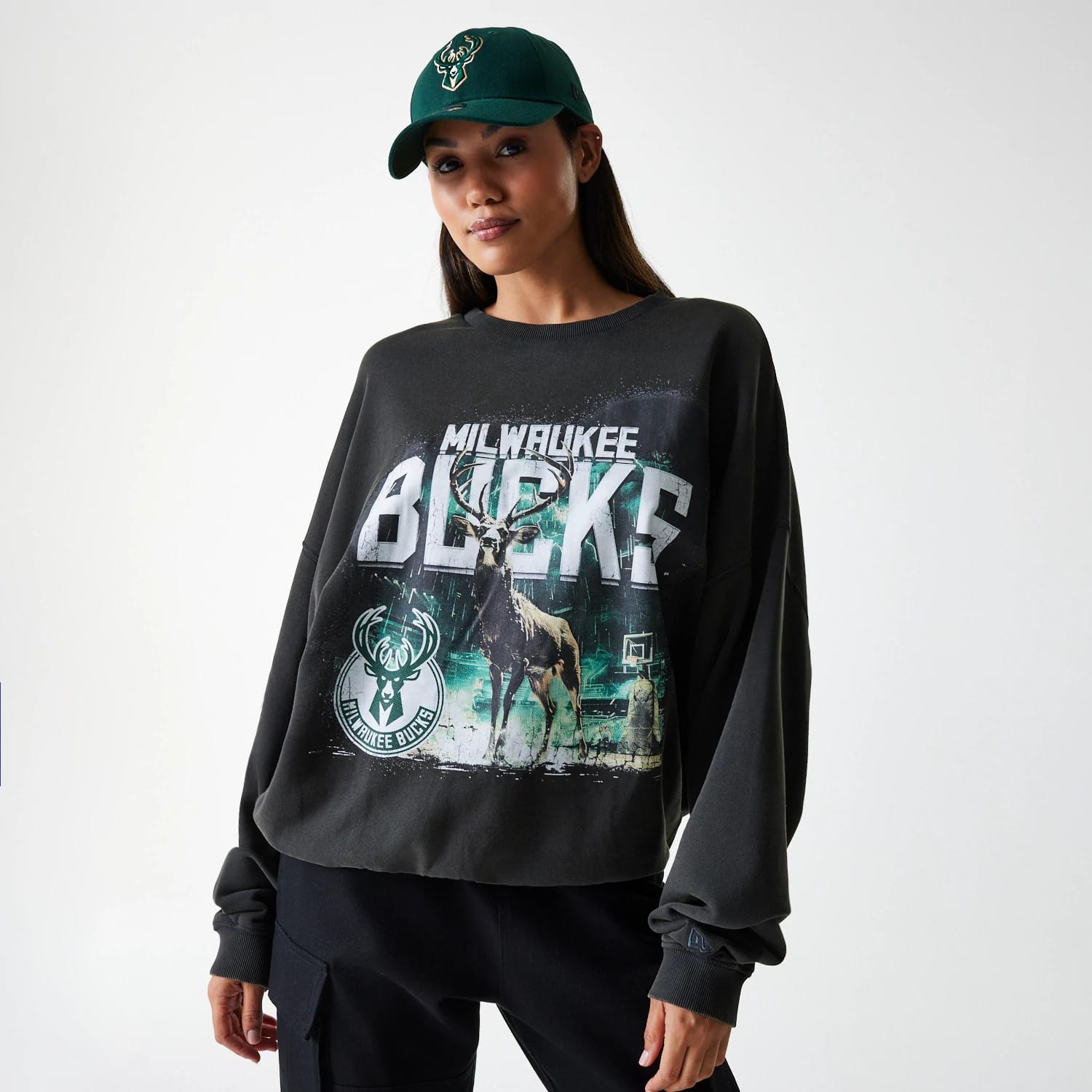 New Era Milwaukee Bucks NBA Washed Graphic Grey Oversized Crew Neck Sweater, on-model showing the front graphic featuring the Milwaukee Bucks name and logo with a prominent deer illustration, paired with a matching dark green cap.