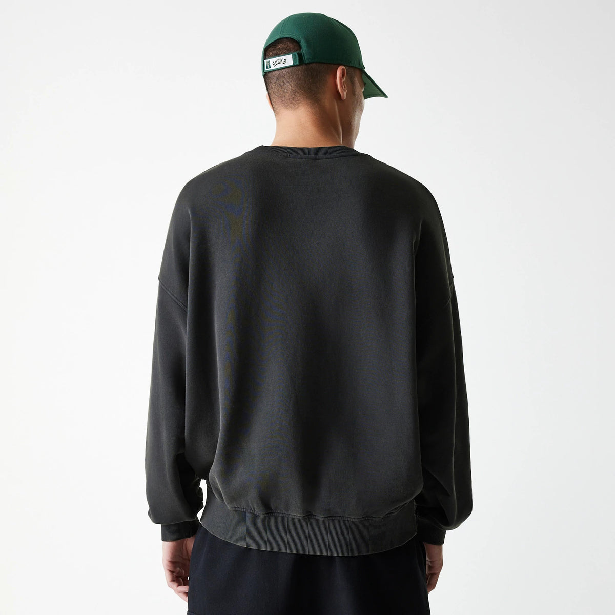 New Era Milwaukee Bucks NBA Washed Graphic Grey Oversized Crew Neck Sweater, back view on a model wearing a green cap, showing the relaxed fit and ribbed hem of the washed grey sweater.