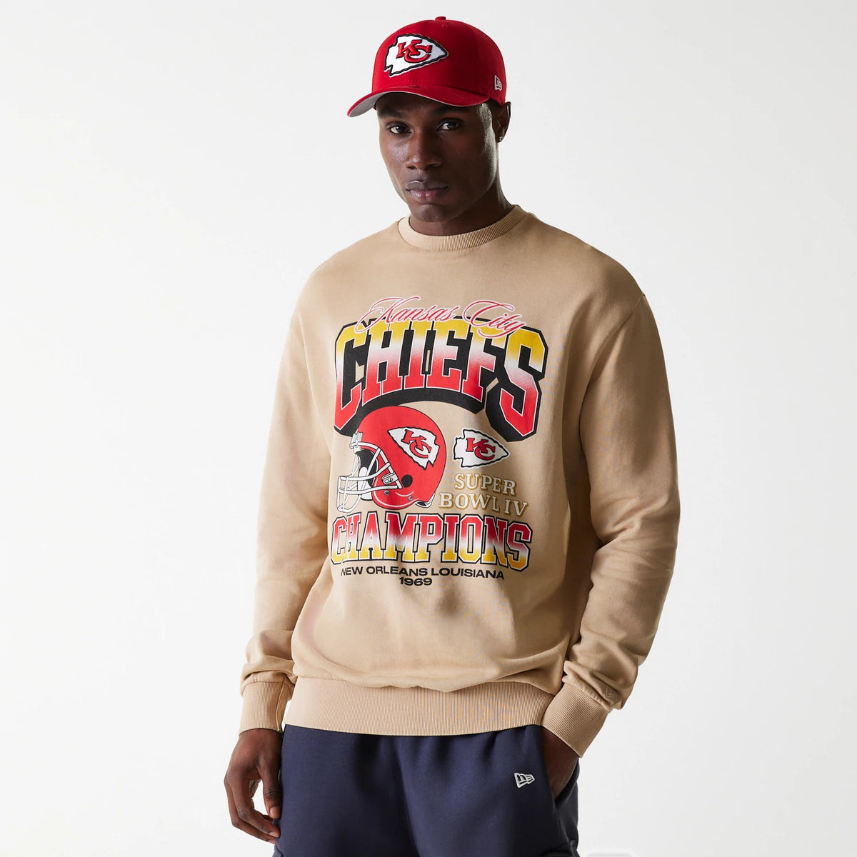 New Era Kansas City Chiefs NFL Washed Super Bowl Beige Oversized Crew Neck Sweater, front view on a model wearing a red cap with the Chiefs logo, featuring large Chiefs lettering and Super Bowl IV Champions graphics on the chest.